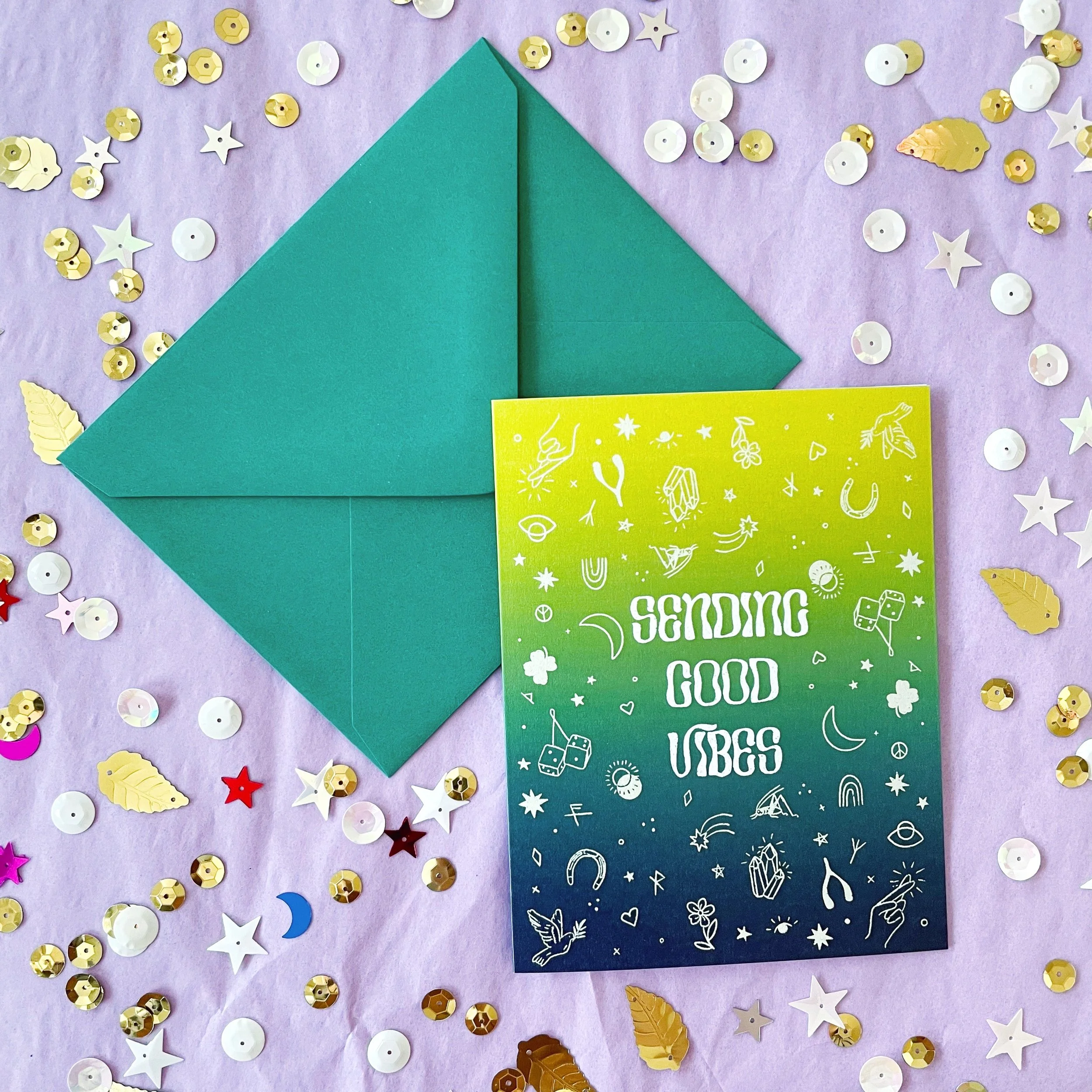 greeting card with drawn good luck charms that says sending good vibes with purple envelope on lavender background with gold and white sequins
