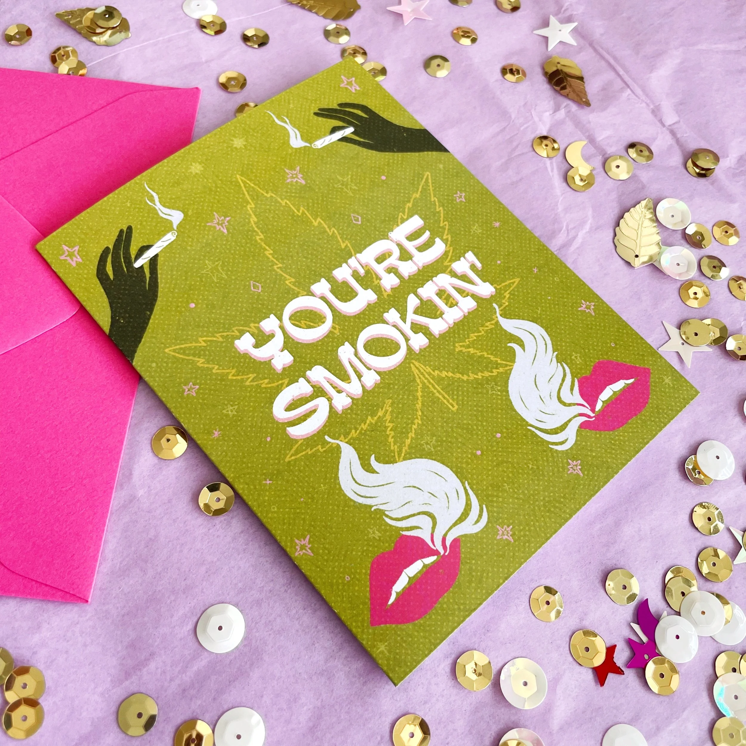 green greeting card that says you're smokin' with cannabis leaf and smoking lips with pink envelope on a lavender background surrounded by gold sequins