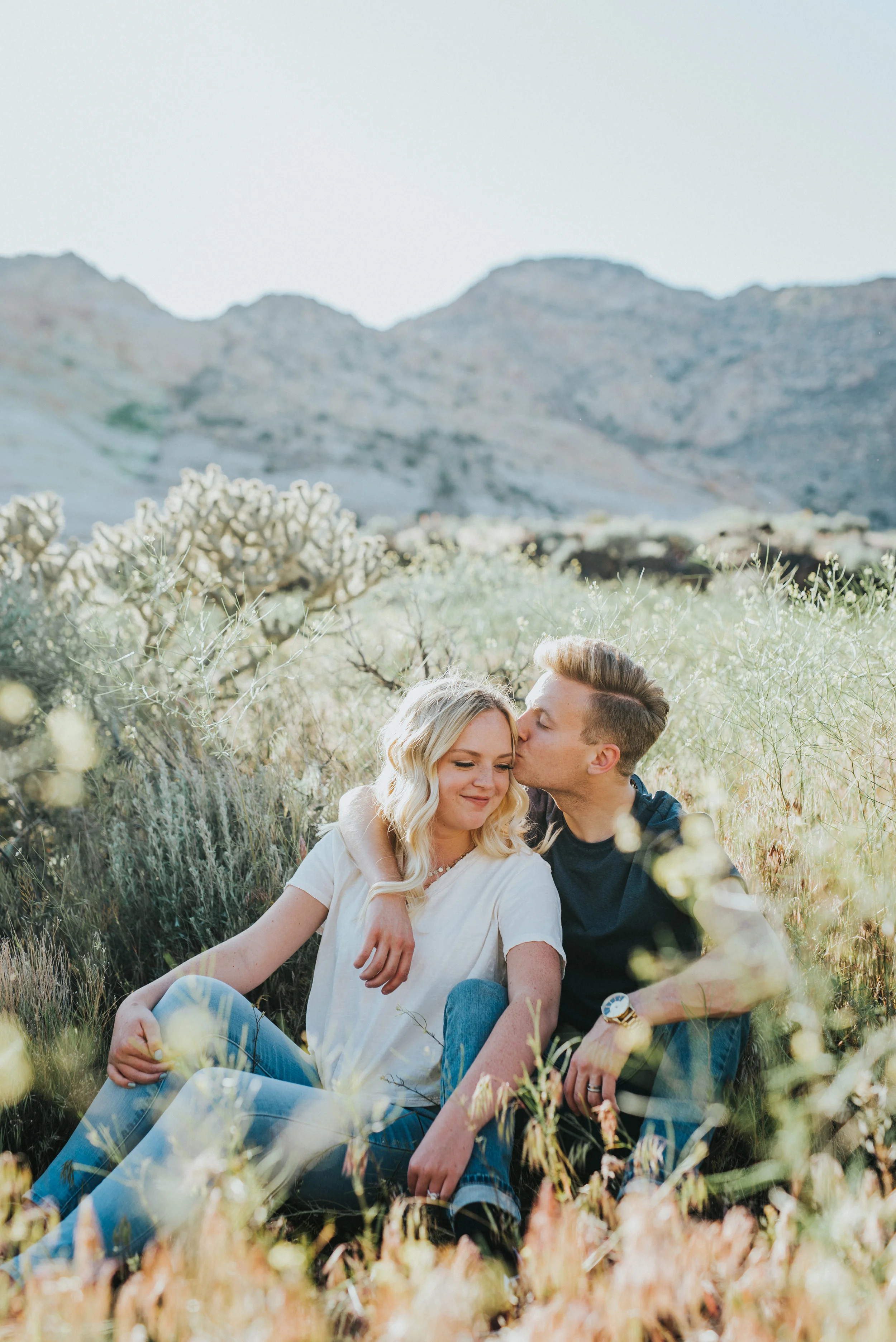  northern utah professional wedding photographer snow canyon outdoor engagement session sitting hugging kissing romantic mountain background tall grasses neutral engagement outfit inspiration #Saintgeorgephotography #snowcanyon #engagementsession #ut