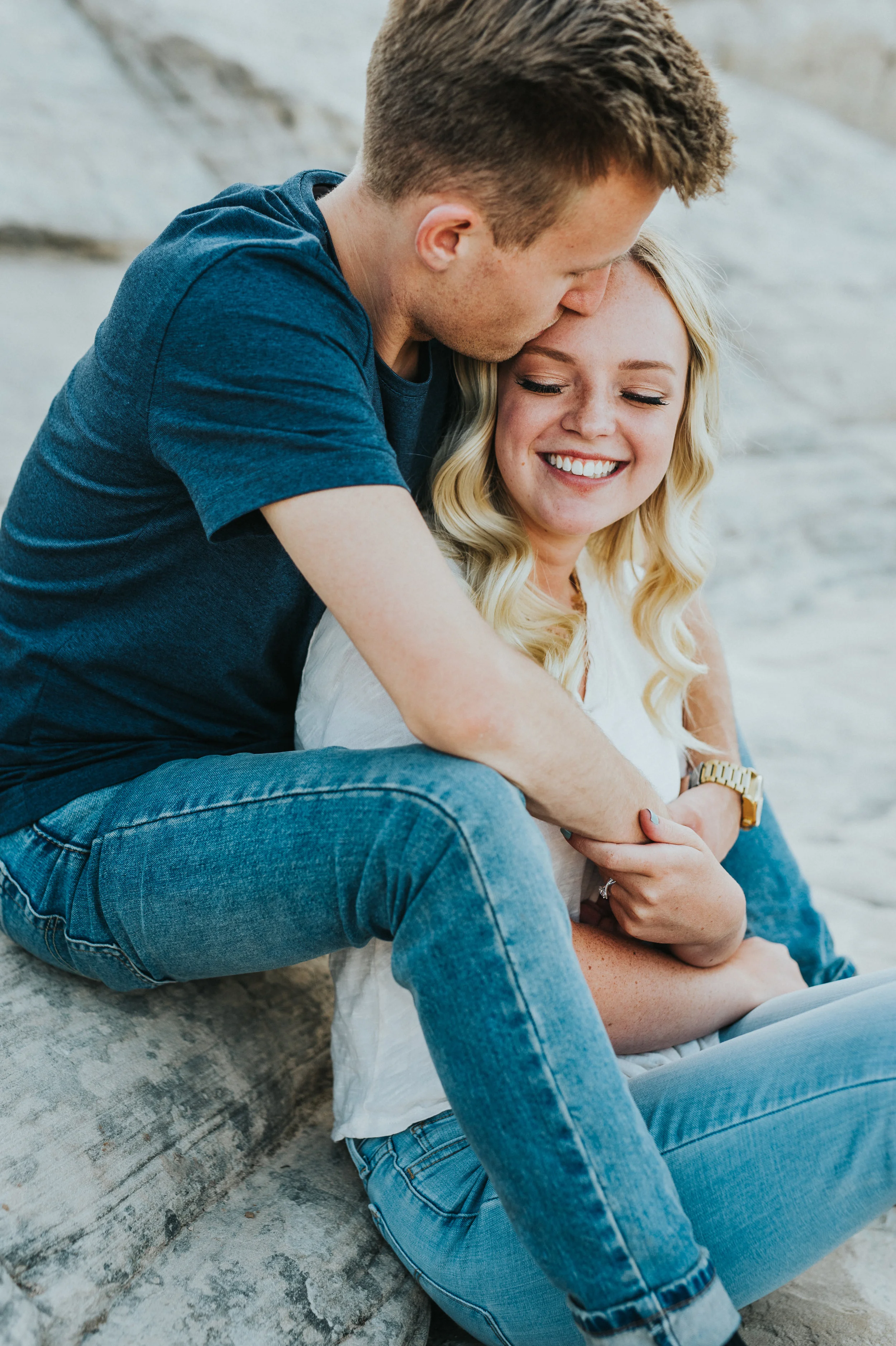  happy looking over shoulder kissing forehead sitting cuddling playful engagement session rocky background mountain outdoor engagement northern utah wedding photography jeans blonde hair #saintgeorgephotography #snowcanyon #engagementsession #utah #h