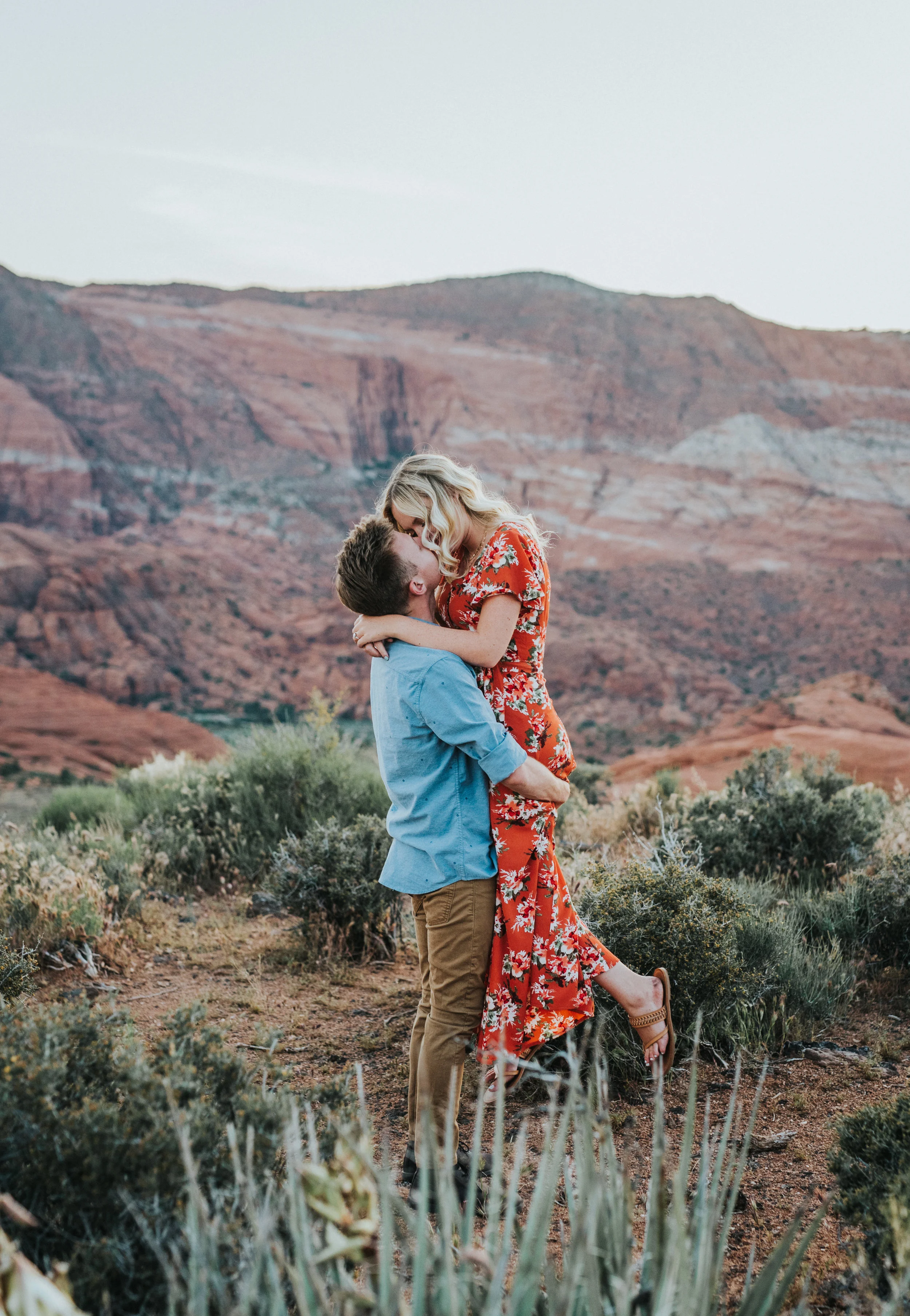  hugging holding jump in arms wild love romantic kissing tall grass red rock snow canyon sunset authentic posing saint george engagement photographer long floral orange dress blonde hair inspiration #saintgeorgephotography #snowcanyon #engagementsess