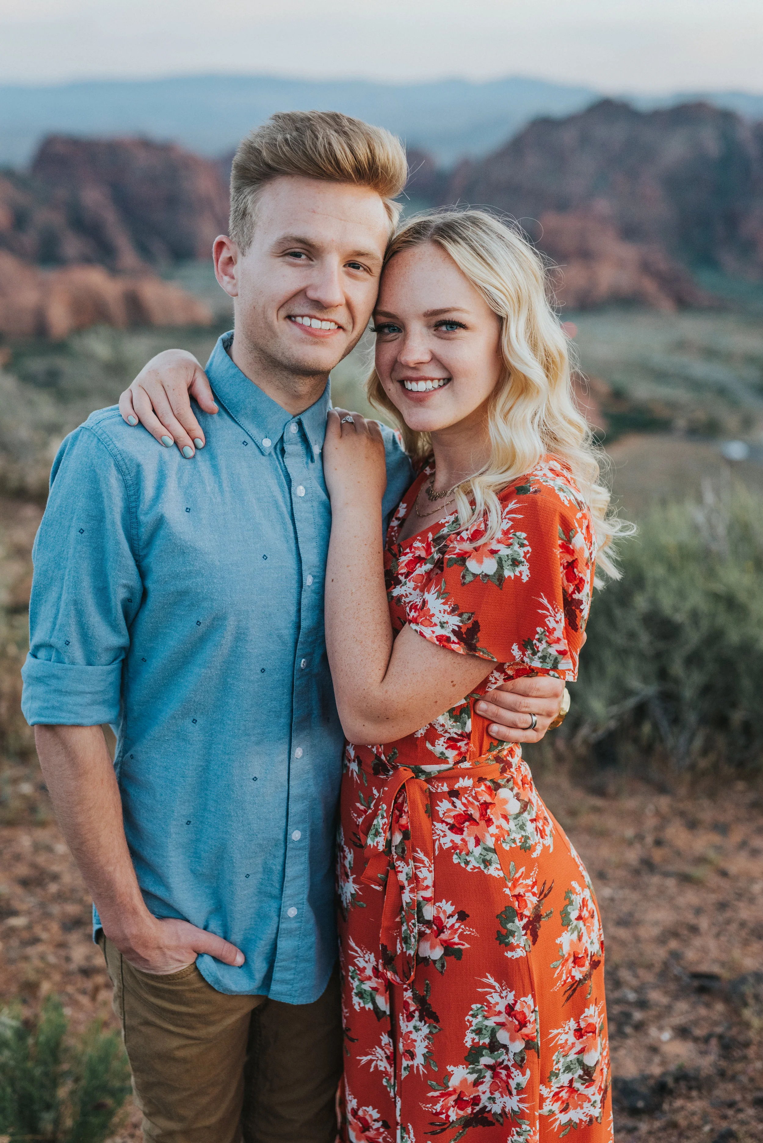  hugging causal posing happy smiling polka dot button up shirt khaki pants burnt orange floral dress blonde wavy hair inspiration red rocks snow canyon saint george photographer #saintgeorgephotography #snowcanyon #engagementsession #utah #hairinspir