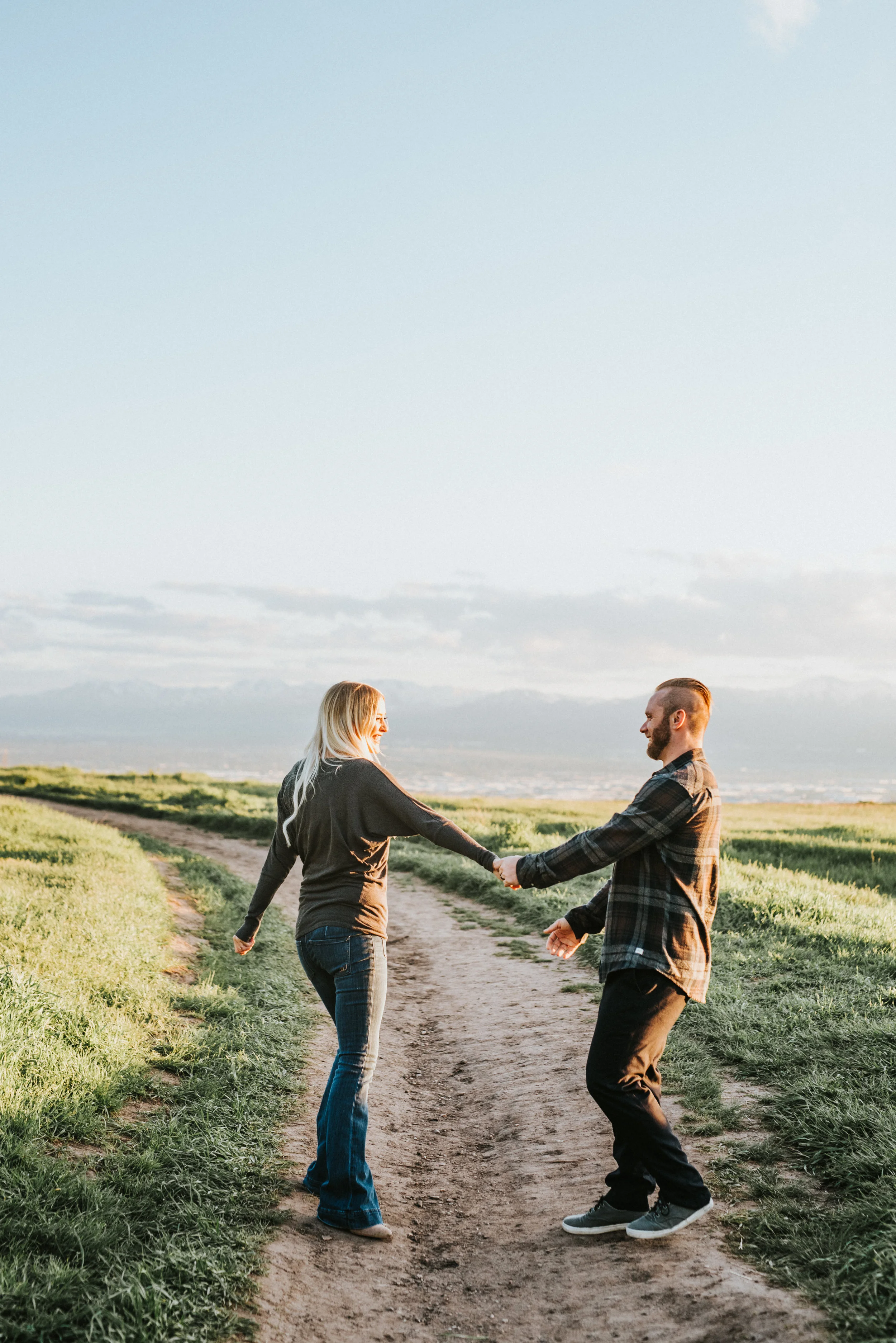  holding hands spinning dirt road path playful laughing engagement session northern utah engagement photographer cache valley runnel springs green field mountain backdrop fall photos neutral colored engagement outfits #engagementphotography #nothernu