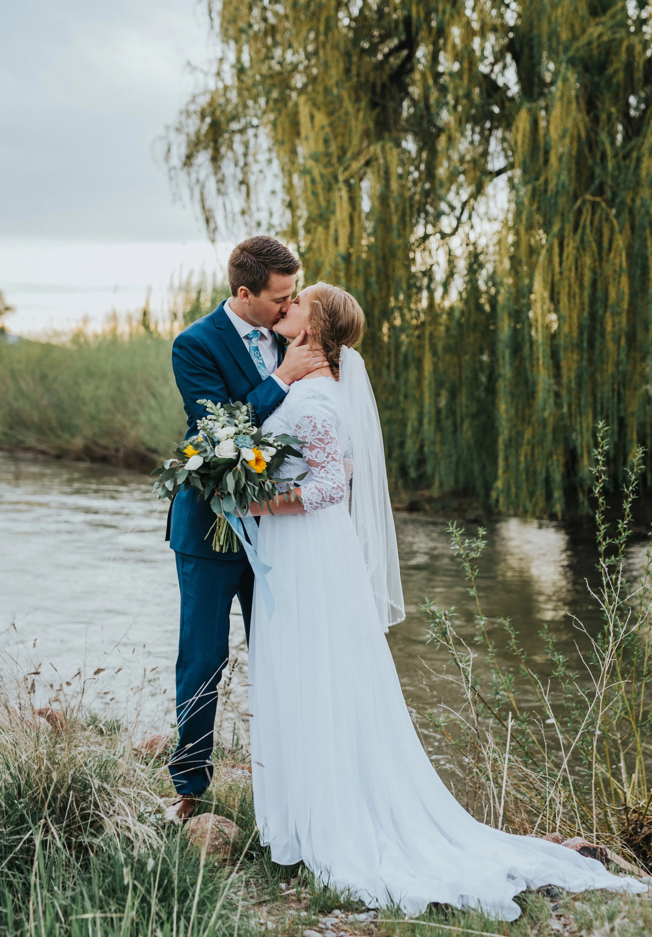  overcast sunset sky willow tree stream tall grass logan utah wedding formals kissing romantic baby blue bouquet ribbon sunflower blue and white floral bouquet modest lace wedding dress cache valley wedding photographer #Loganutah #cachevalleyphotogr