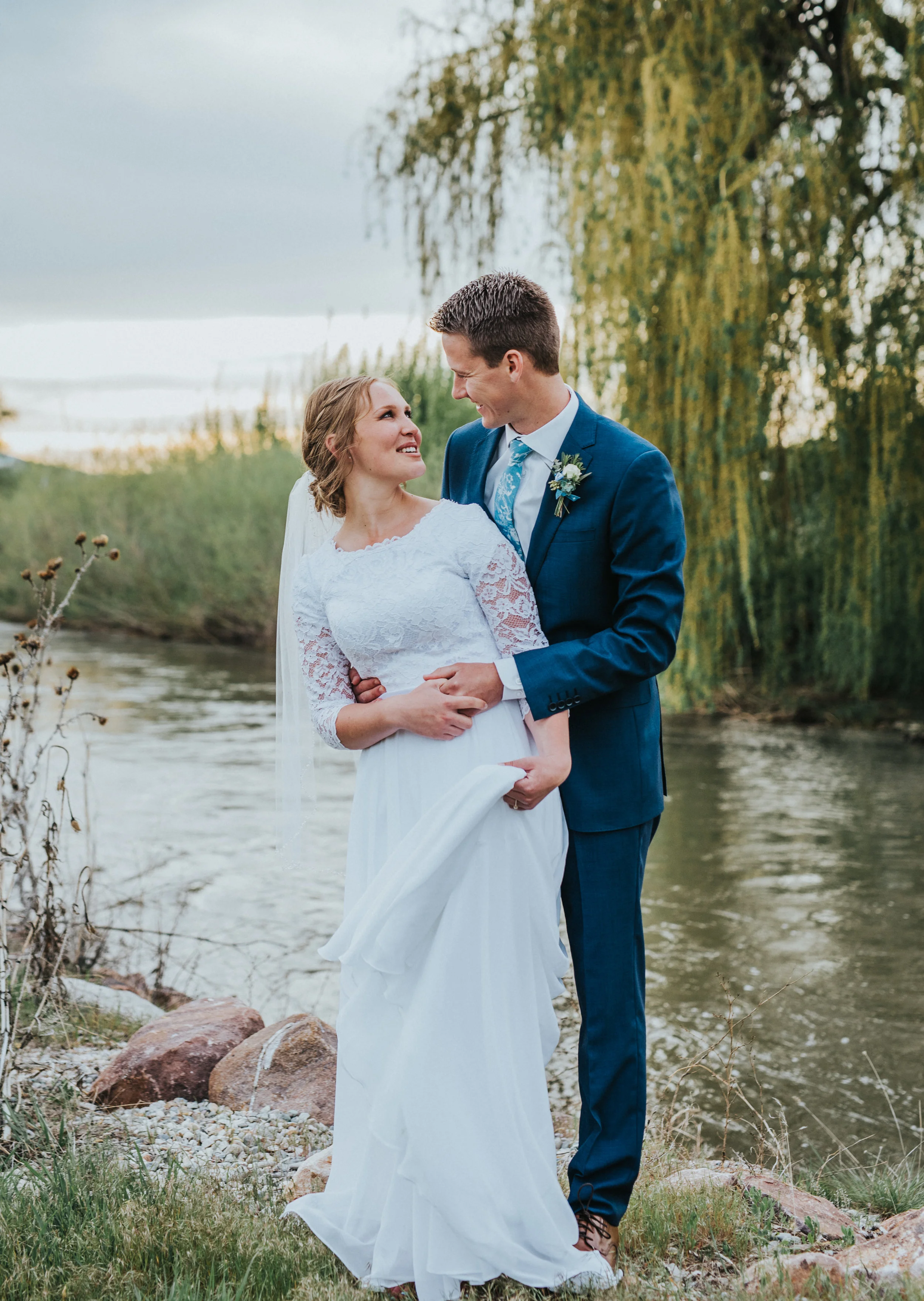  sunset over water overcast skies rocky stream shore logan utah wedding portrait locations hugging hand holding smiling romantic messy braided wedding updo professional cache valley wedding photographer #Loganutah #cachevalleyphotographer #Ido #Isaid