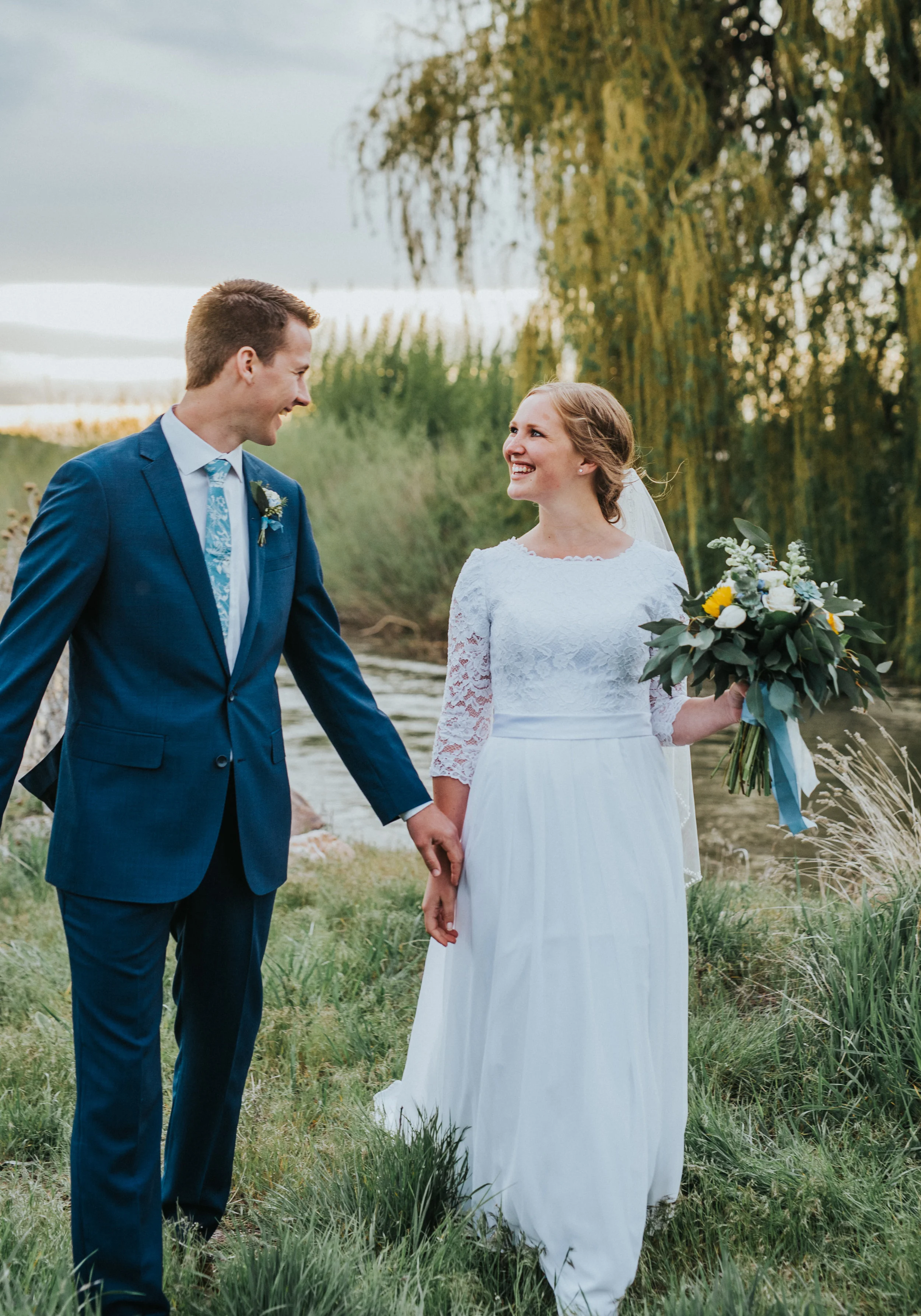  stream bank walking holding hands smiling overgrown grass willow tree logan utah photography locations sunflower blue wedding bouquet wedding dress modest blue wedding suit cache valley wedding photographer #Loganutah #cachevalleyphotographer #Ido #