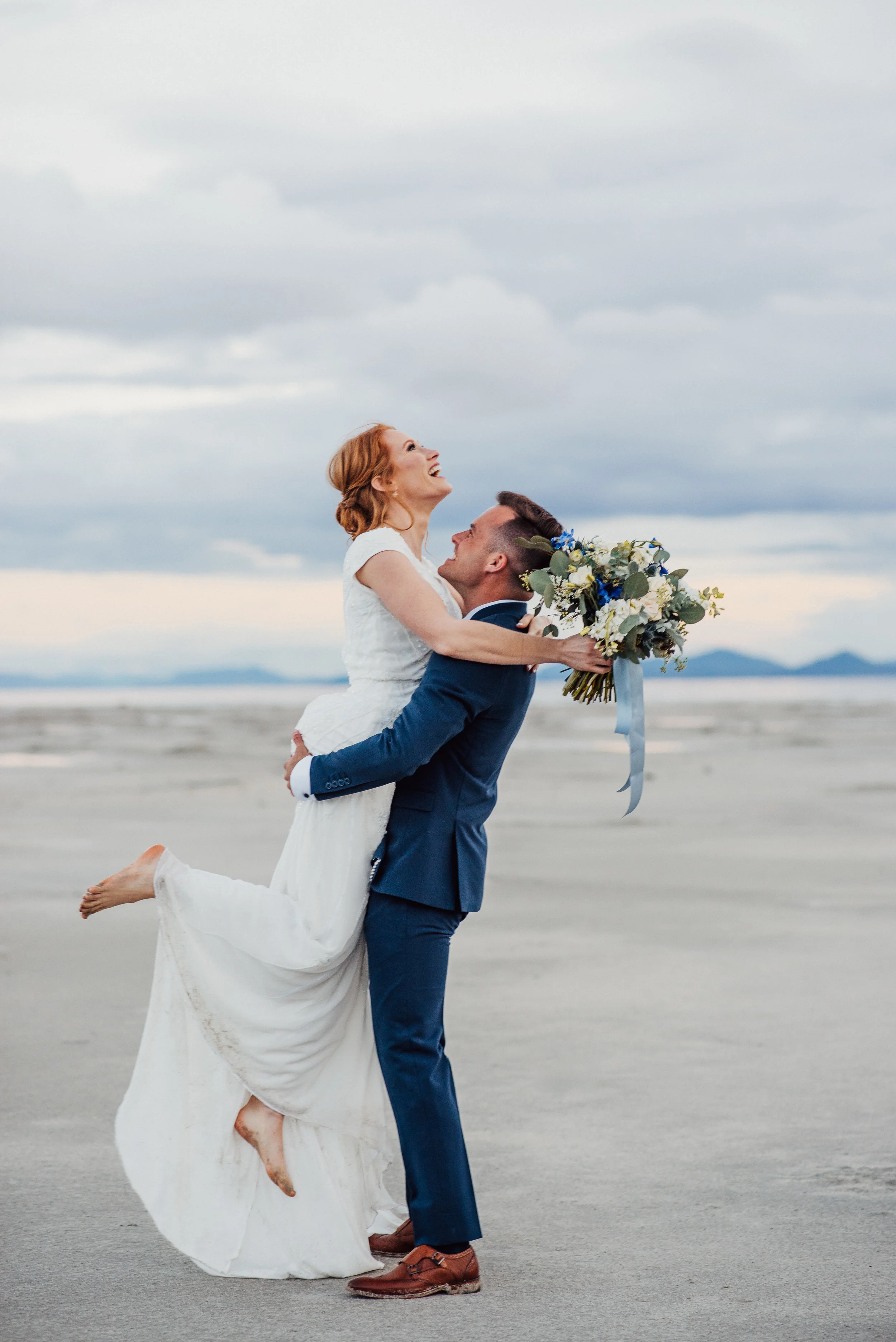 hugging kissing laughing groom picks up bride neutral floral bouquet the great salt lake professional northern utah photographer playful authentic bridal session #spiraljetty #saltlakecityweddingphotographer #saltlake #ido #northernutahphotographer 