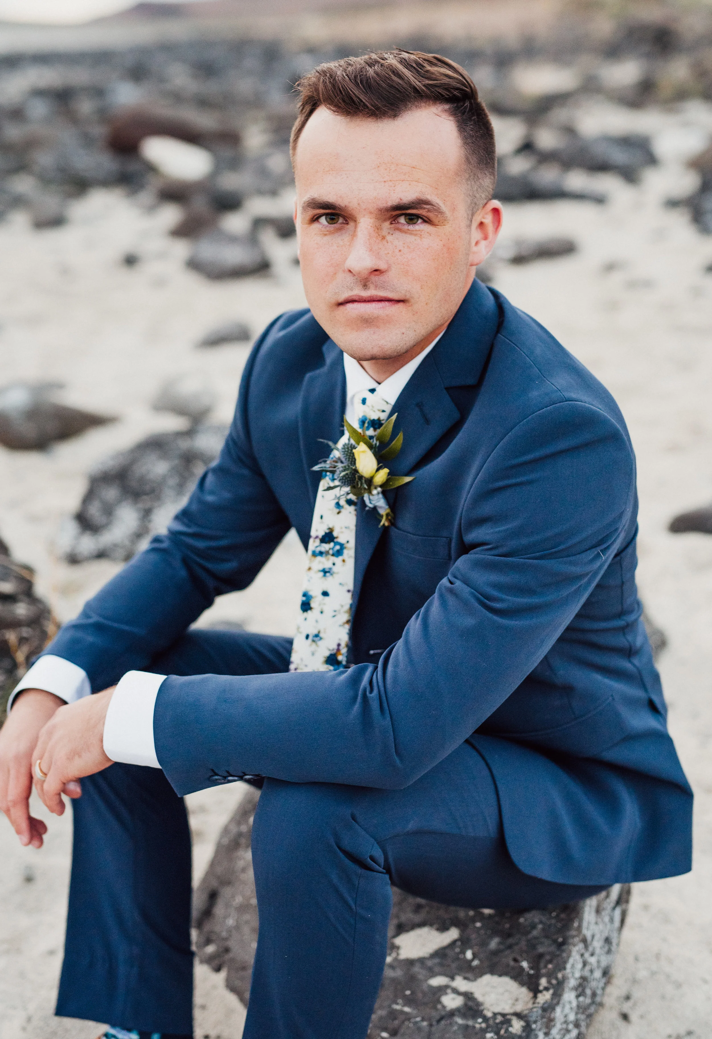  groom navy wedding suit sitting on rocks sandy ground bridal photos floral tie northern utah wedding photographer sunset bridal session #spiraljetty #saltlakecityweddingphotographer #saltlake #ido #northernutahphotographer #bridals #weddinginsp #hai
