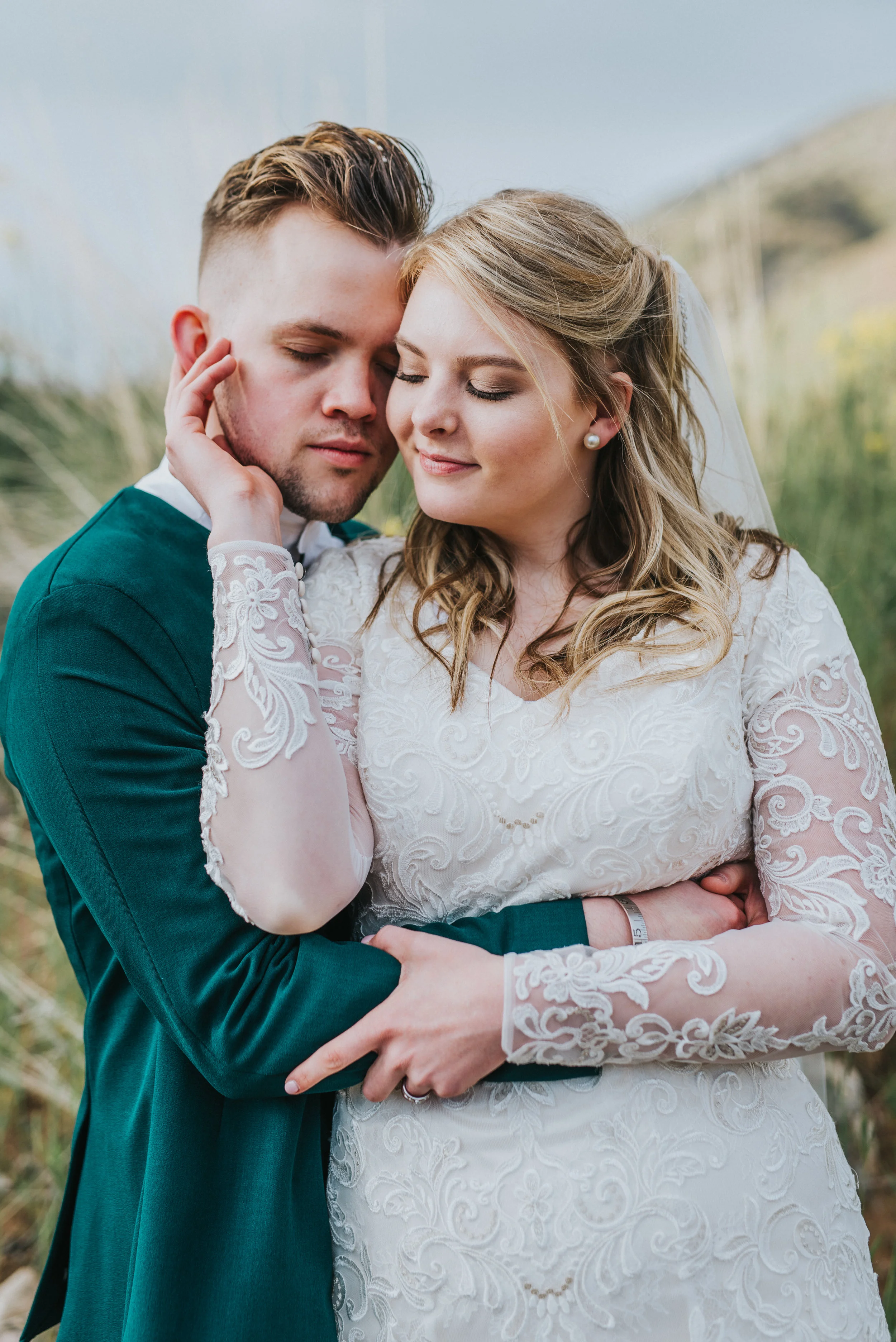  standing hugging cupping face bridal portraits hyrum utah black smith fork reservoir logan utah norther utah wedding photographer emerald green suit jacket modest lace wedding dress tall grass mountains #cachevalleyutah #hyrumutah #northernutahphoto