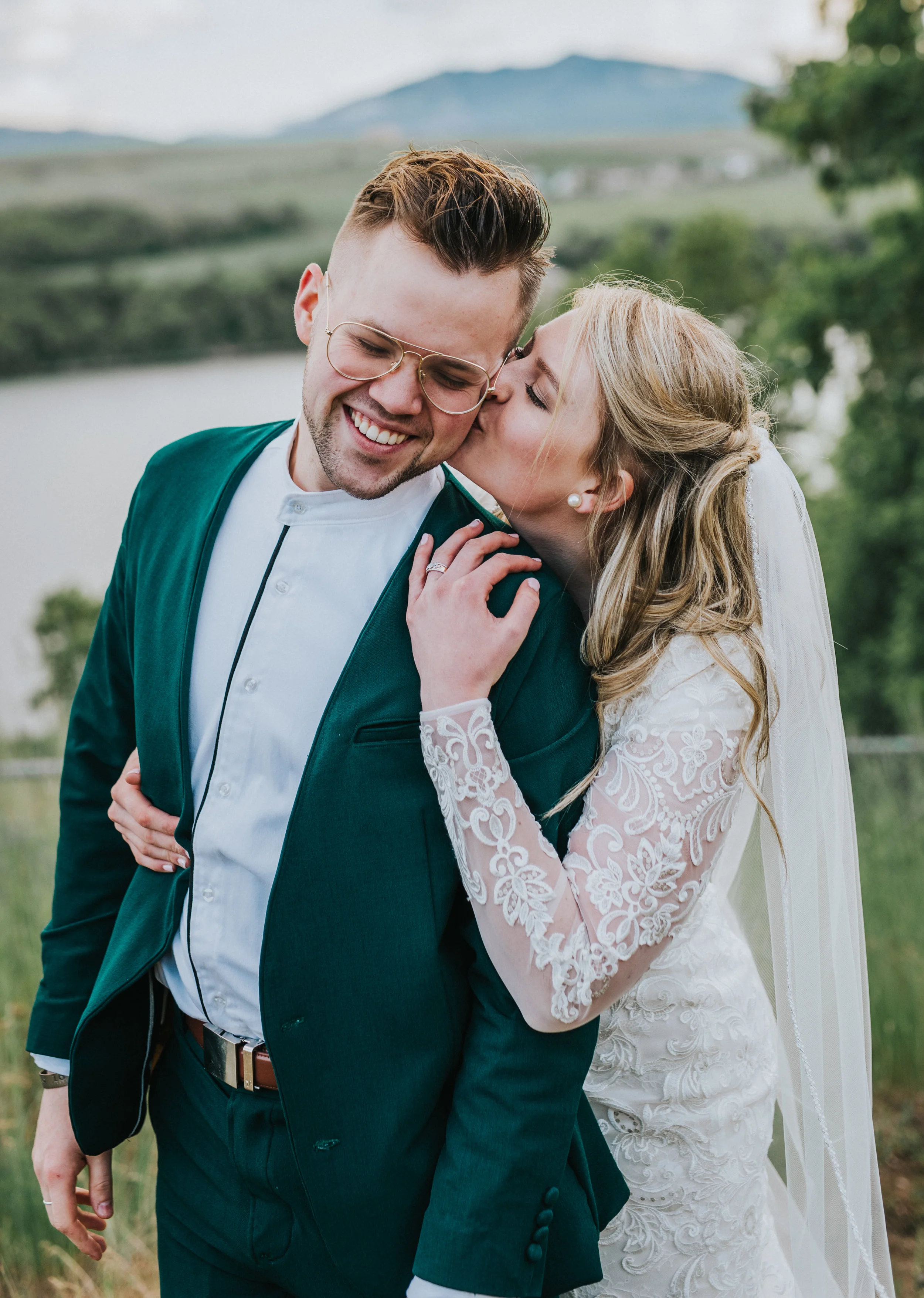  kissing cheeks laughing smiling playful wedding portraits emerald green wedding suit lace wedding dress modest loose blonde curls long veil reservoir water mountains logan utah northern utah wedding photographer #cachevalleyutah #hyrumutah #northern
