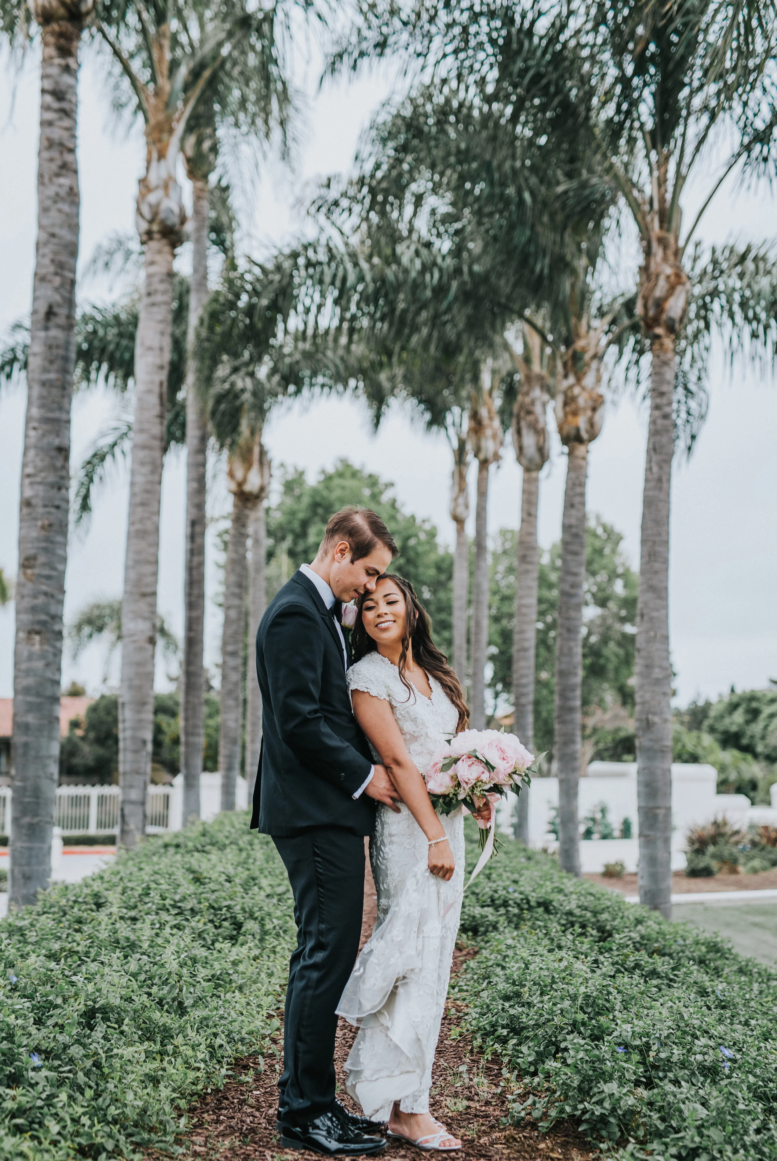  plam tree temple grounds san diego temple wedding lds hugging smiling moody skies modest beaded wedding dress black wedding suit bow tie pink peony wedding bouquet boutonniere northern utah wedding photographer san diego wedding photography #lajolla