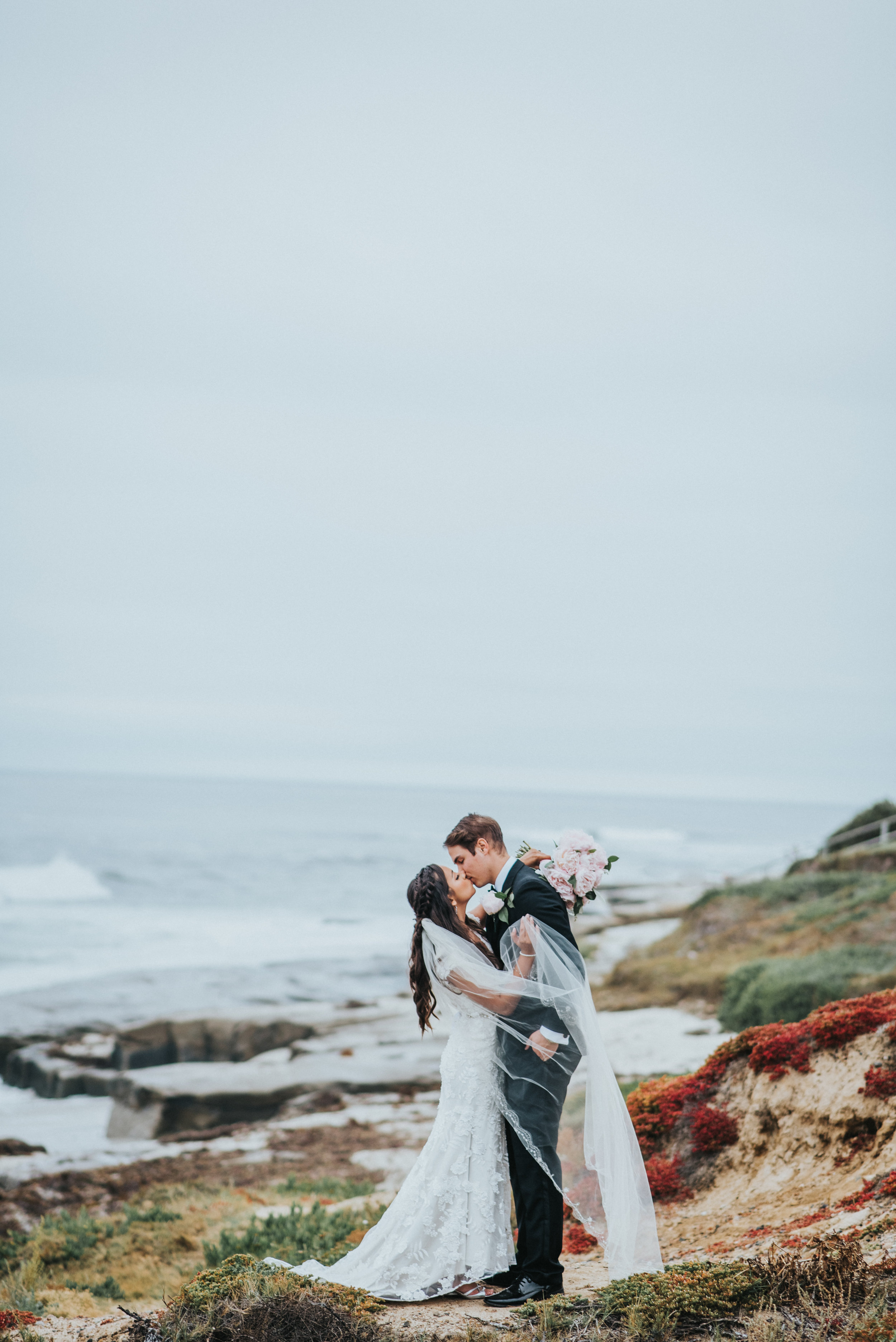  moody skies romantic kissing cliffs billowy long bridal veil hugging peony bridal bouquet hugging windansea beach la jolla california northern utah wedding photographer #lajollacaliforniaweddingphotographer #sandiegotemple #windanseabeachcalifornia 