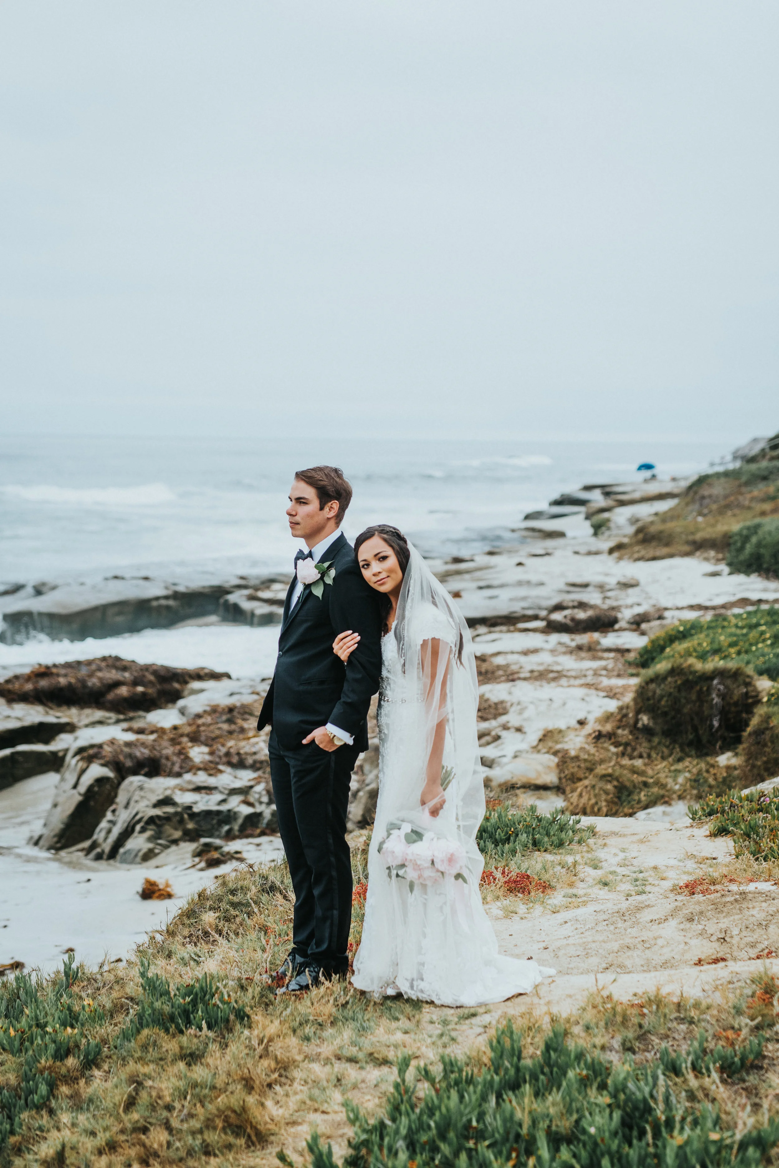  wedding day beach photos moody ocean skies cliffside bridals head on shoulder hugging romantic lace bridal dress san diego wedding photographer groom hands in pockets #lajollacaliforniaweddingphotographer #sandiegotemple #windanseabeachcalifornia #n