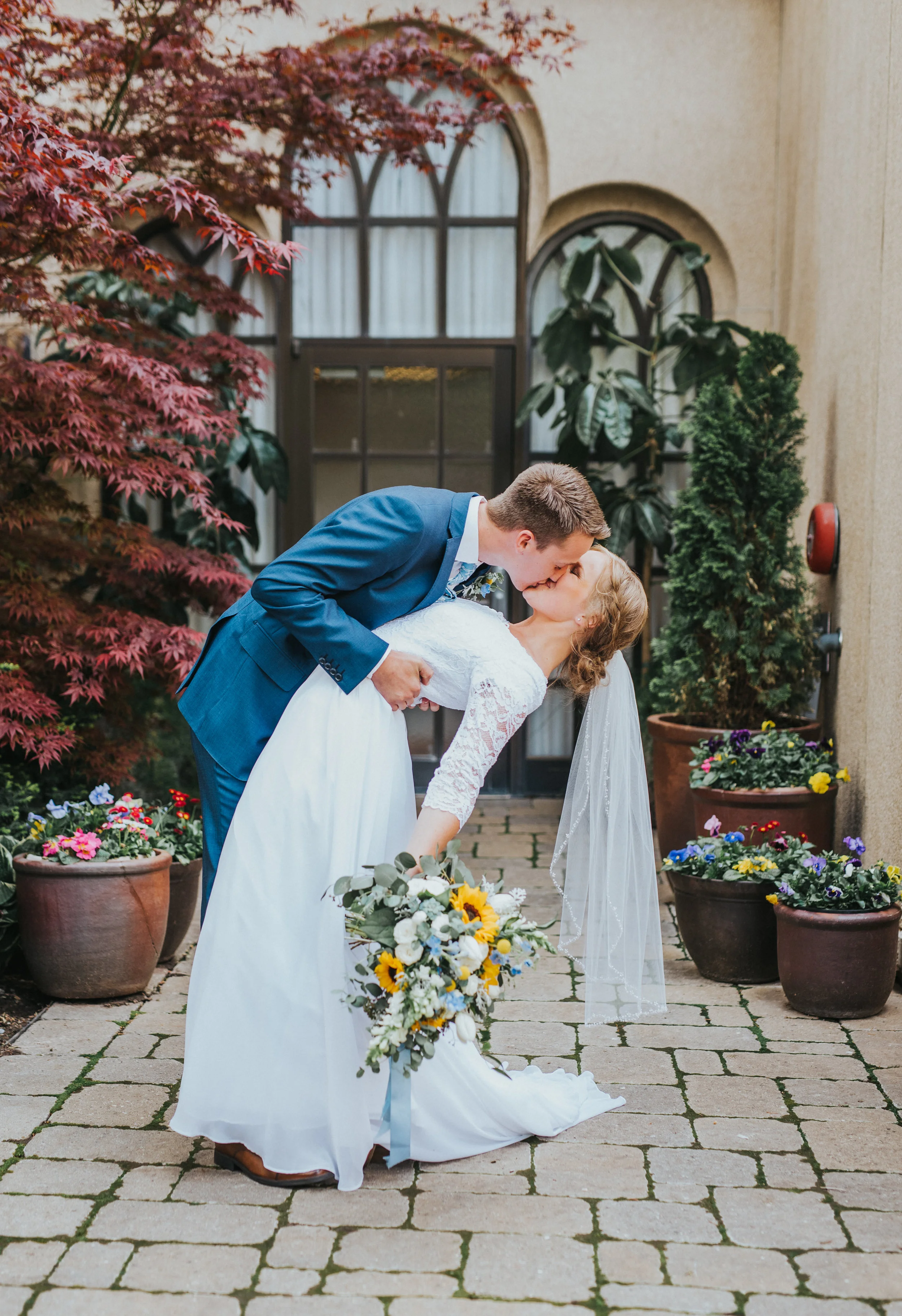  just married romantic kissing hugging dipping modest lace wedding dress lace sleeve blue wedding suit braided updo courtyard floral wedding bouquet yellow flowers dasies professional logan utah wedding photographer #Loganutah #cachevalleyphotographe