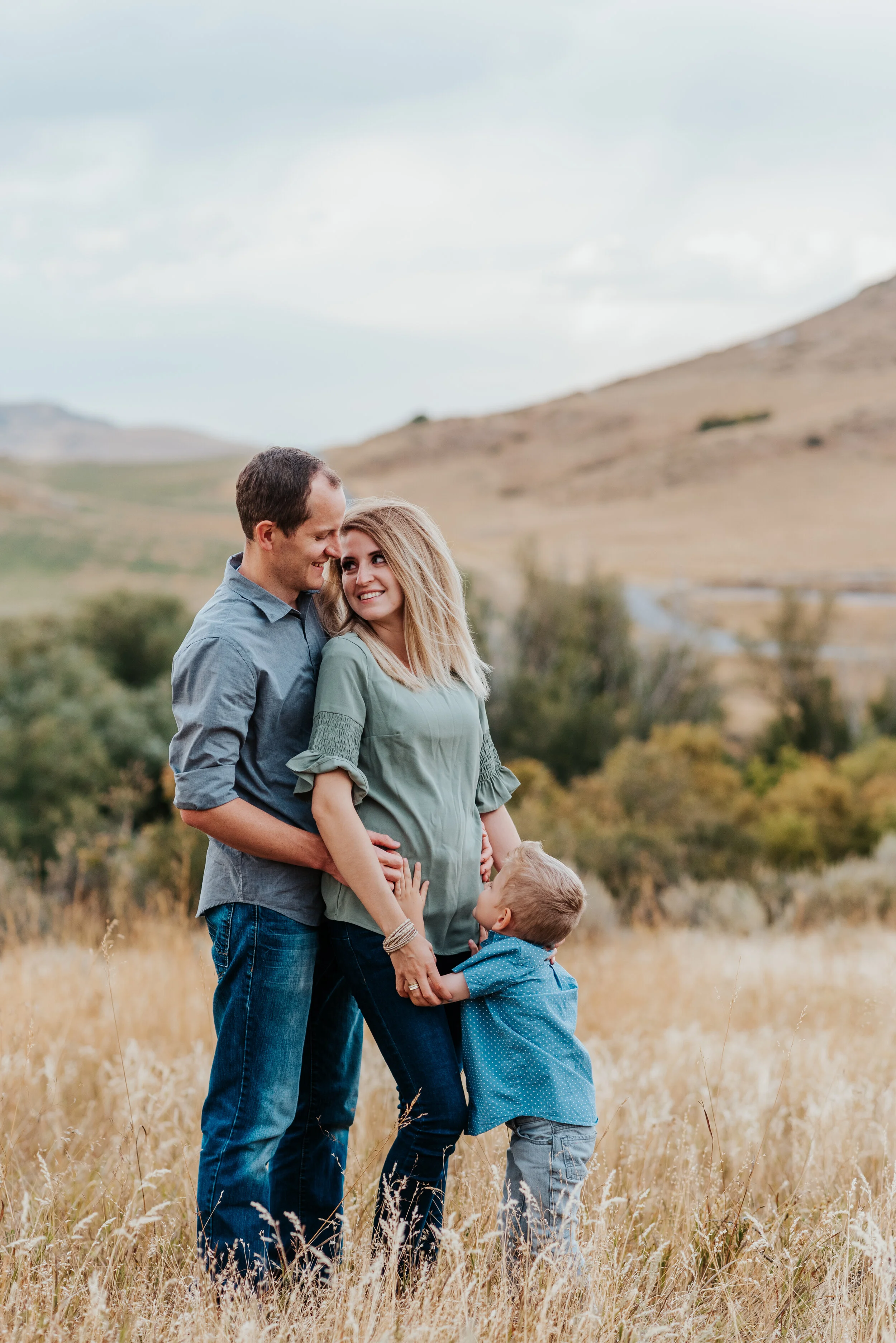  Husband and son hugging their wife and mother in the tall golden grassy field. sweet parent and children poses baby announcement in family portraits in logan utah cache valley photographers in northern utah professional photographers in utah family 