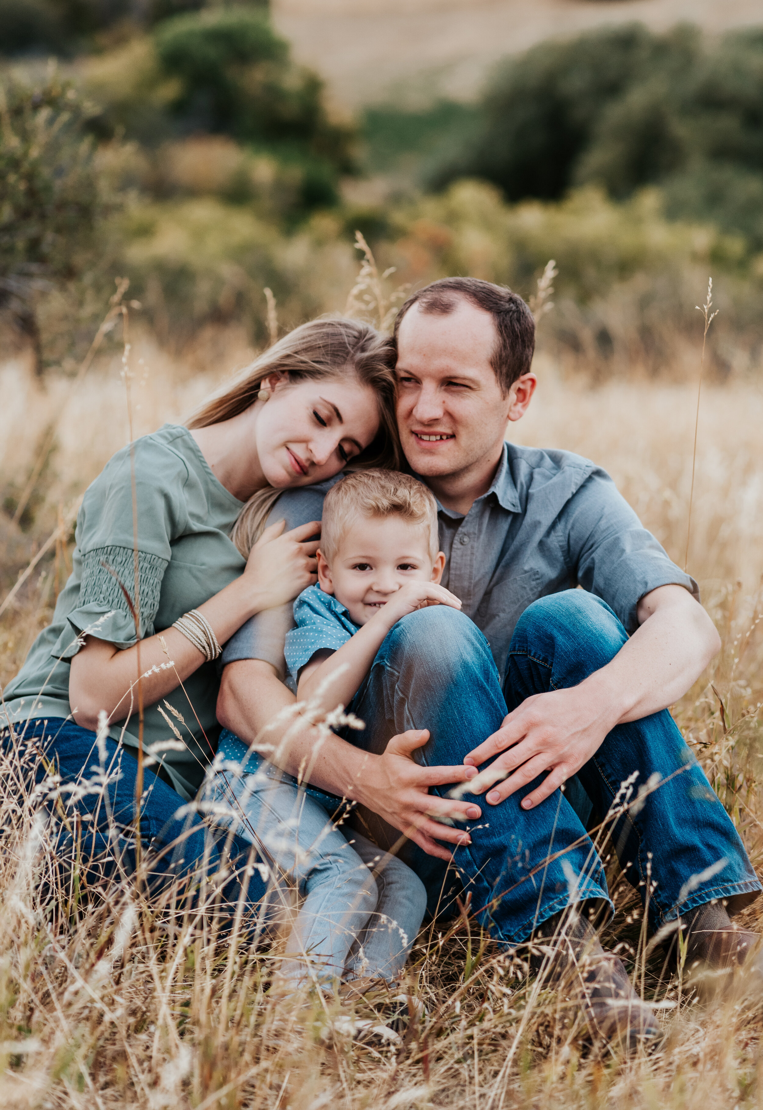  Family of three cuddled in a field in the mountains near Logan, Utah. casual outfit styles sweet parent and children poses baby announcement in family portraits in logan utah cache valley photographers in northern utah professional photographers in 