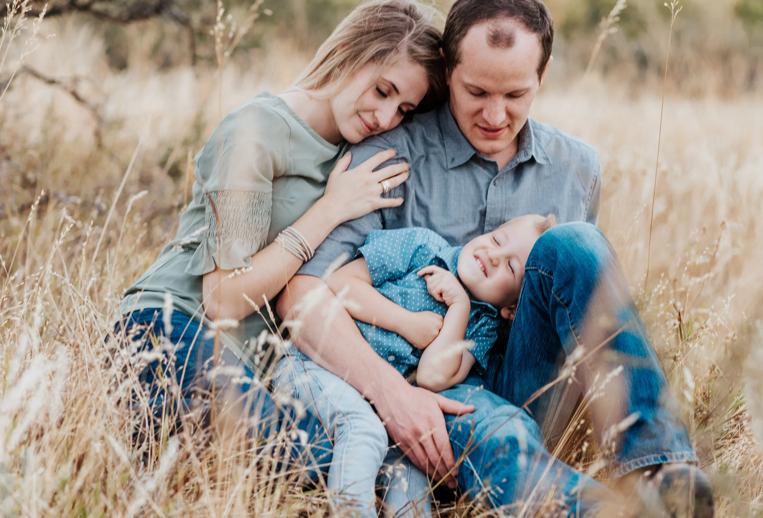 Happy parents cuddling with their cute son in a field of tall grass. baby announcement in family portraits in logan utah cache valley photographers in northern utah professional photographers in utah family pose inspiration casual outfit styles swee…