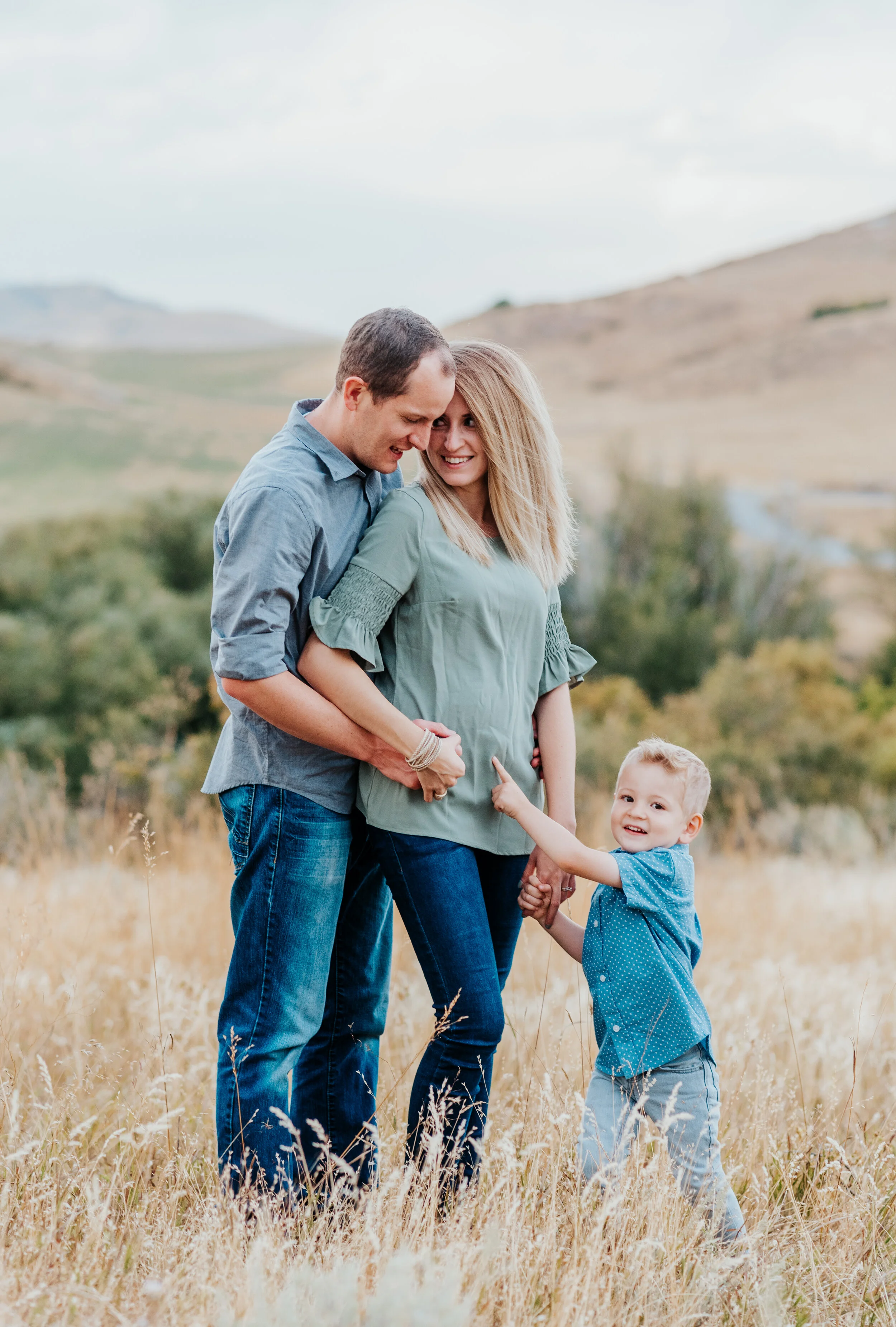  Mom and Dad holding her baby bump while son points to the baby sister in his mom’s belly. family pregnancy pose inspiration casual outfit styles sweet parent and children poses baby announcement in family portraits in logan utah cache valley photogr