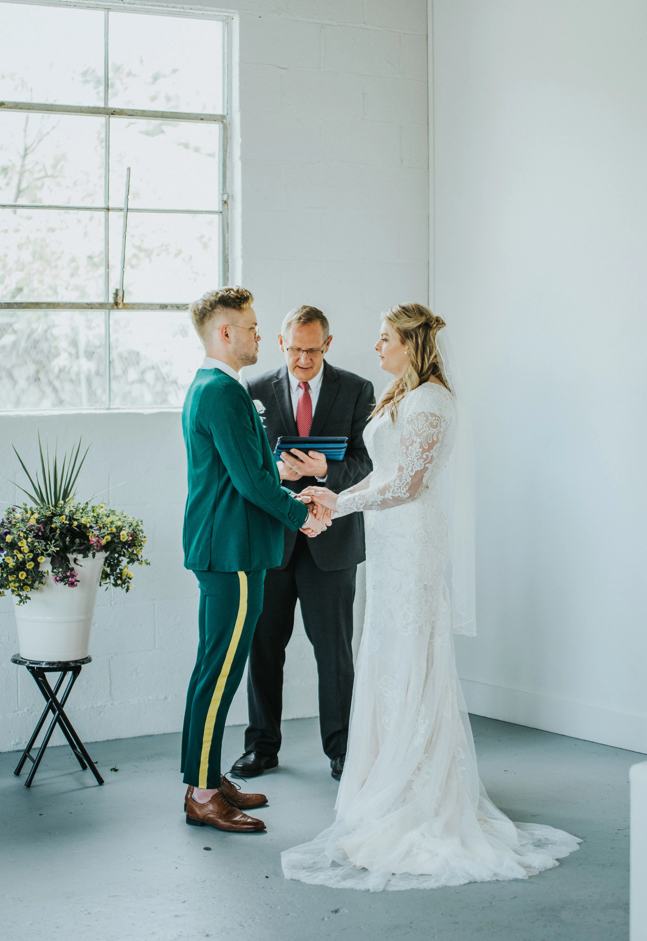  white studio wedding ceremony holding hands i do loose wedding curls retro wedding suit emerald green suit yellow striped pant officiant modest white lace wedding dress long sleeved mid length wedding veil professional logan utah wedding photographe