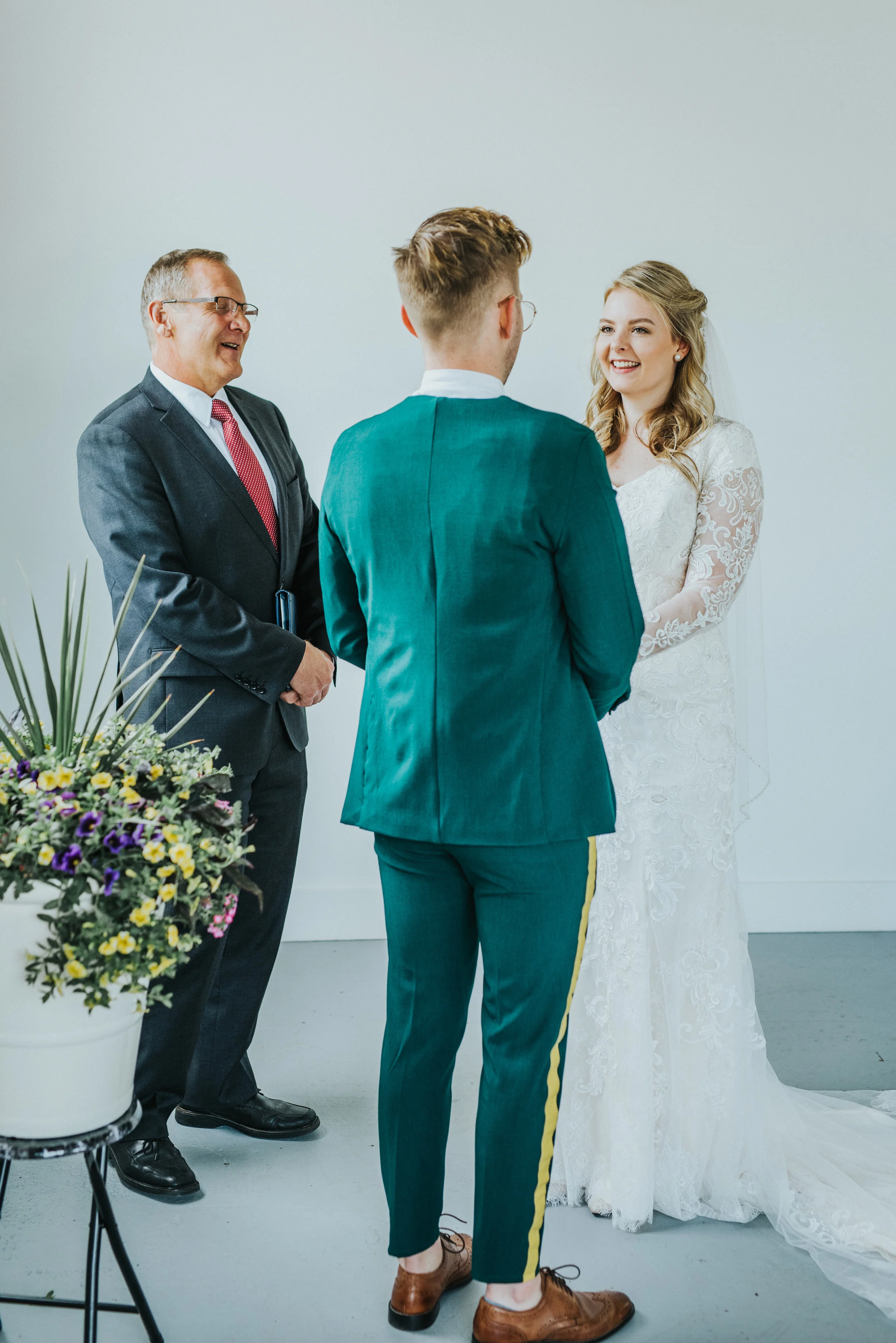  emerald green wedding suit yellow striped pant mens haircut holding hands smiling white studio wedding ceremony officiant bride groom wedding inspiration brown mens dress shoe whote long sleeved lace wedding dress professional wedding photographer l