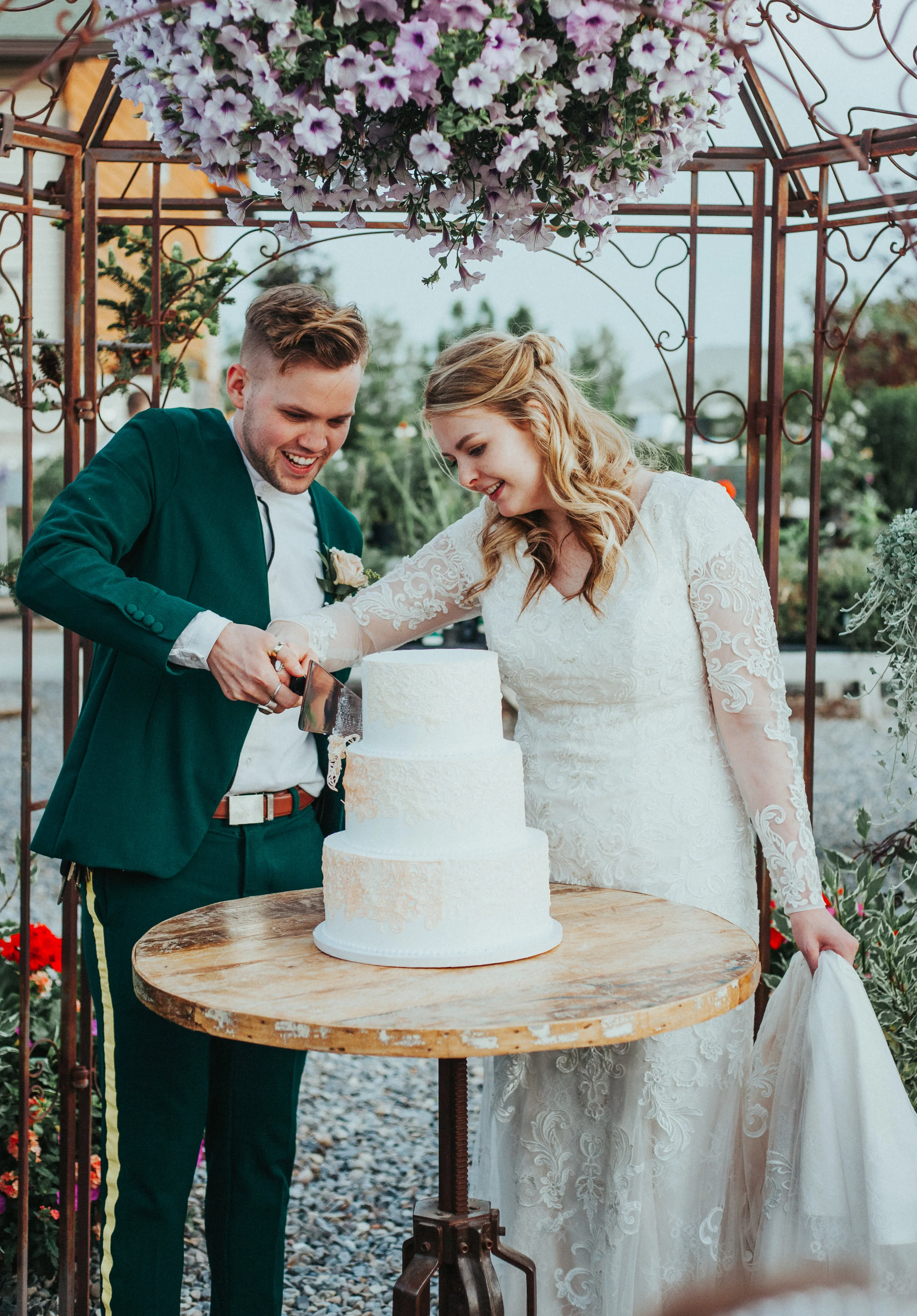  three tiered ivory white lace wedding cake cutting cake half updo wedding hair holding train holding hands retro green wedding suit vintage mens glasses floral archway freckle farm outdoor wedding reception professional logan utah wedding photograph
