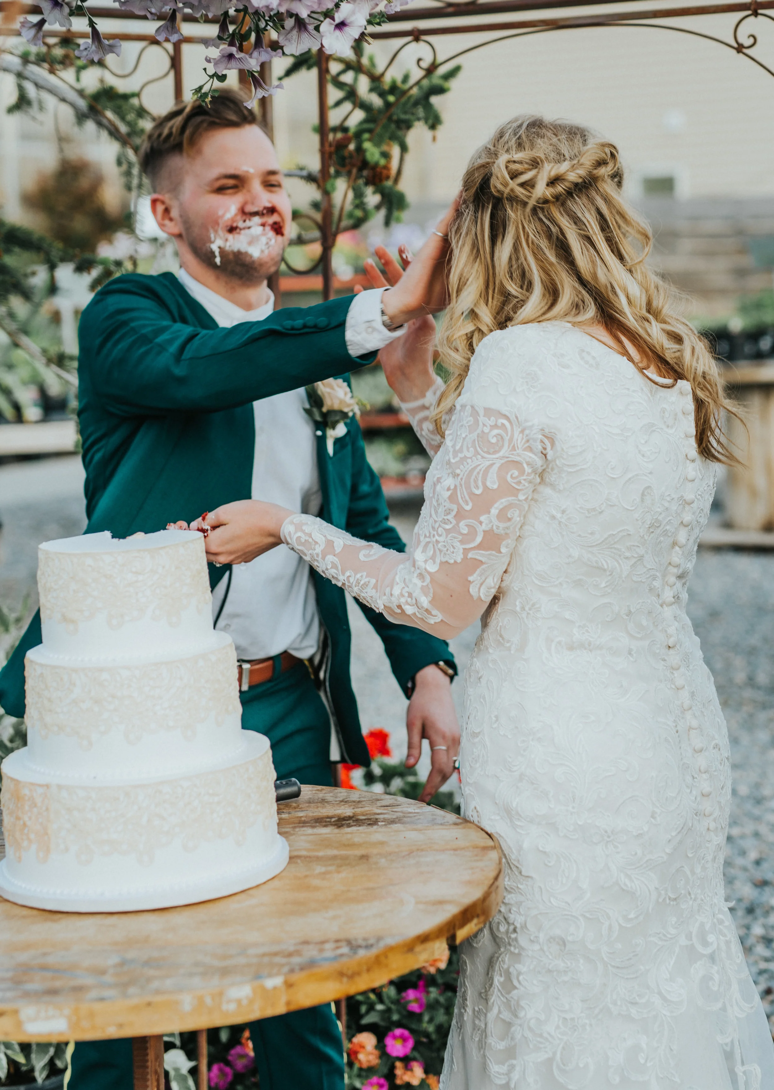   three tiered ivory lace wedding cake smashing cake messy faces happy playful outdoor wedding reception freckle farm reception professional logan utah wedding photographer floral archway holding train modest long sleeved lace wedding dress #StationS
