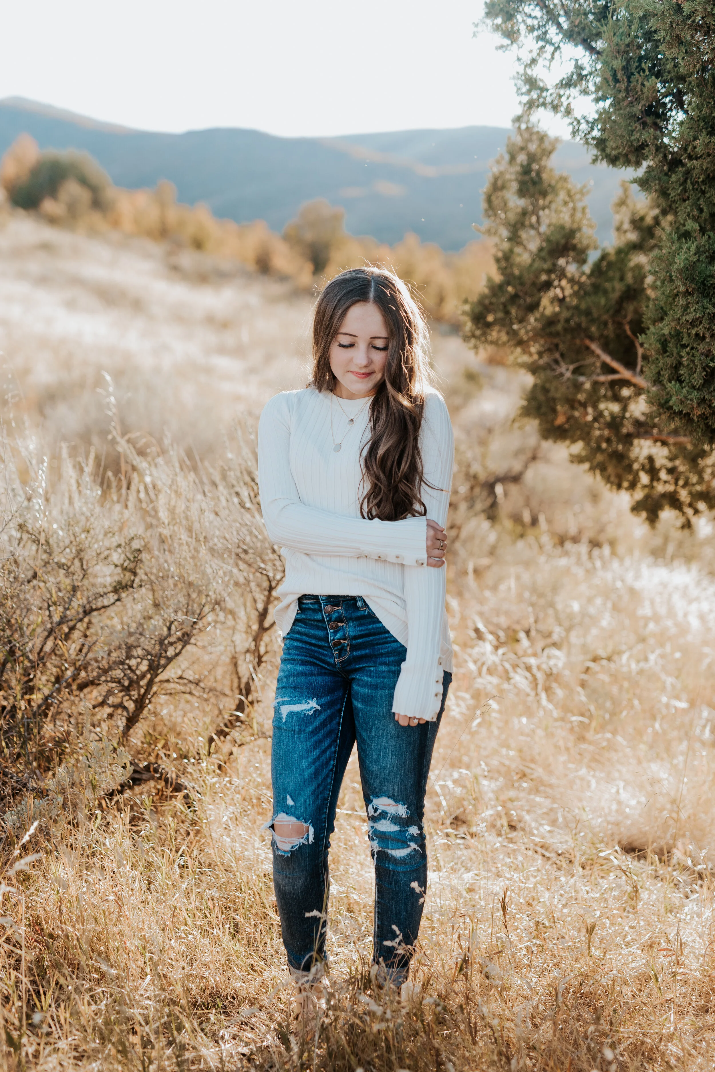  High school girl with long wavy brunette hair wearing a white sweater and dark wash denim in a golden field. teen fashion senior pictures photo session outfit inspiration casual senior picture outfits senior in high school graduating class of 2020 p