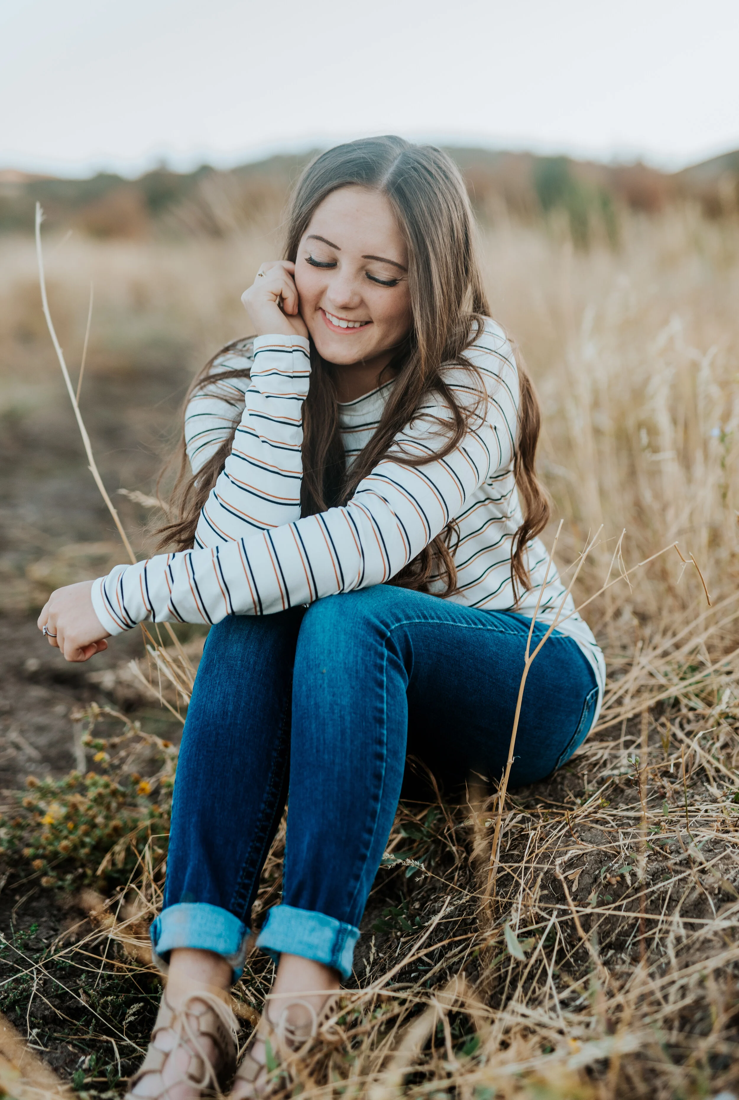  Beautiful teenage girl in a striped long sleeve shirt and dark jeans sitting in a field. logan utah photographers teen fashion senior pictures photo session outfit inspiration casual senior picture outfits senior in high school graduating class of 2