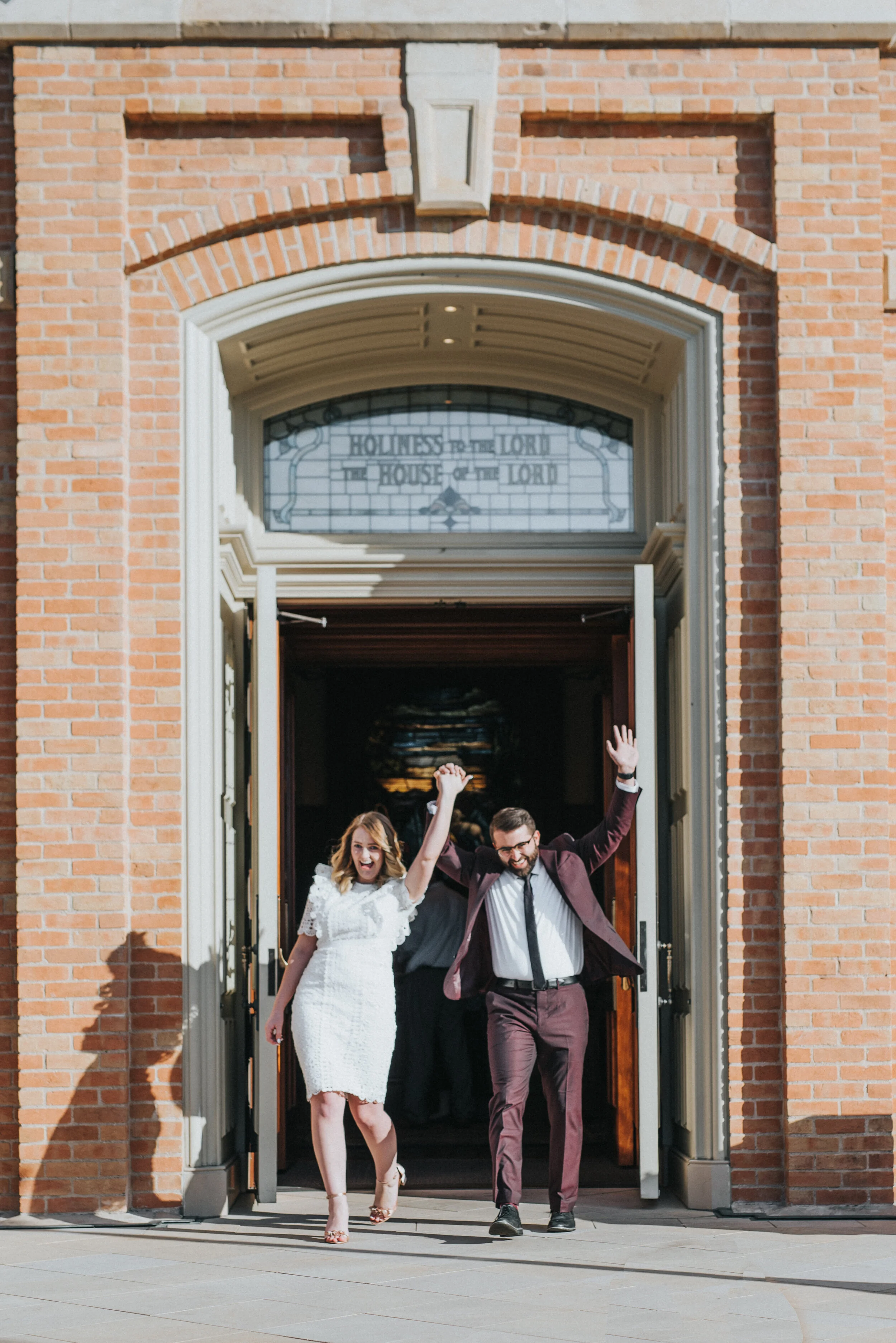  provo city center temple exit celebration hands in the air smiling happy burgundy grooms suit knee length lace wedding dress professional wedding photographer utah valley provo utah holding hands just married #utahvalleyweddingphotographer #provopho
