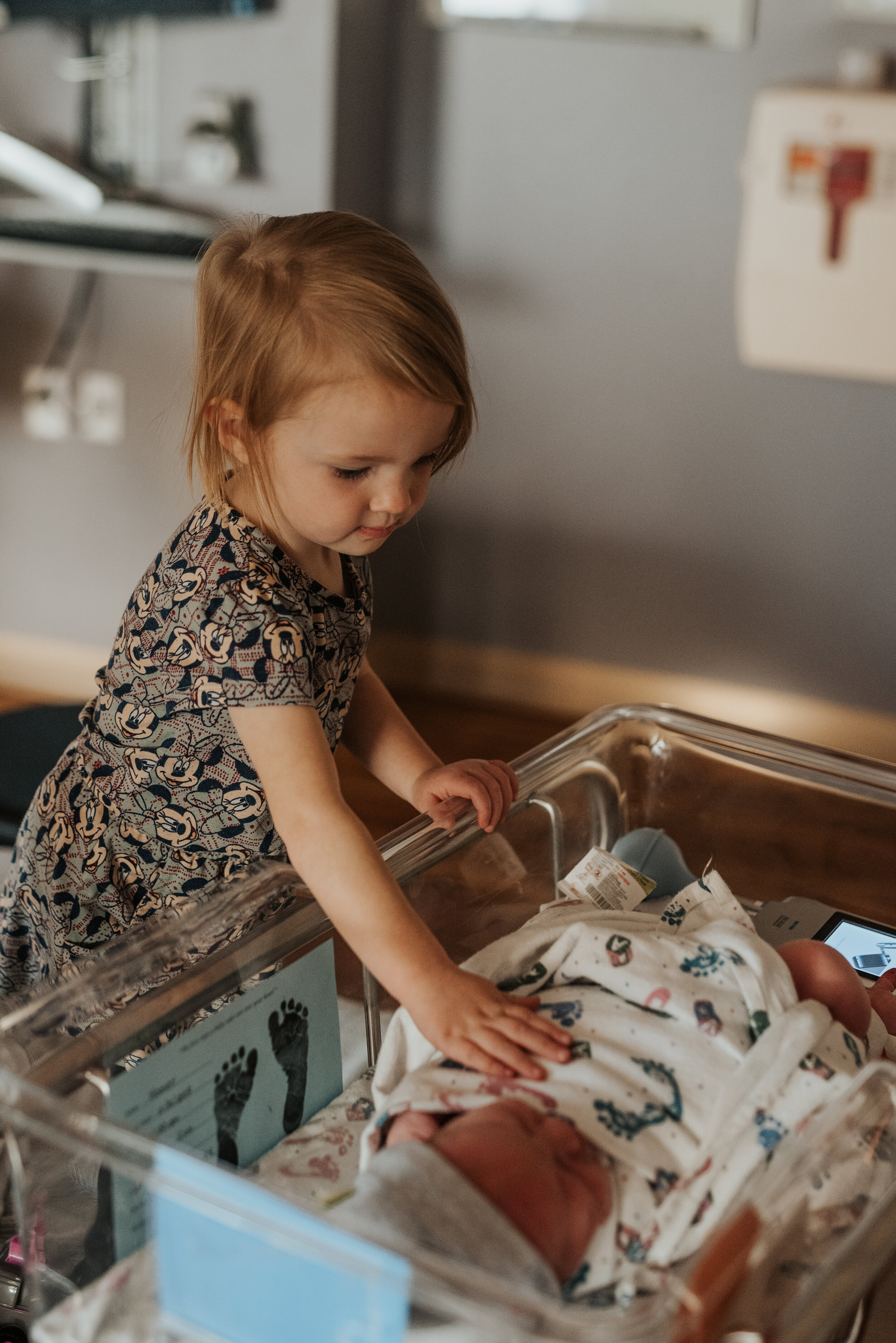  Older sister lovingly resting her hand on her newest little brother in the hospital bassinet. sibling love newborn photographer in cache valley logan utah professional birth story photographer hospital pictures of a brand new baby birth photo sessio