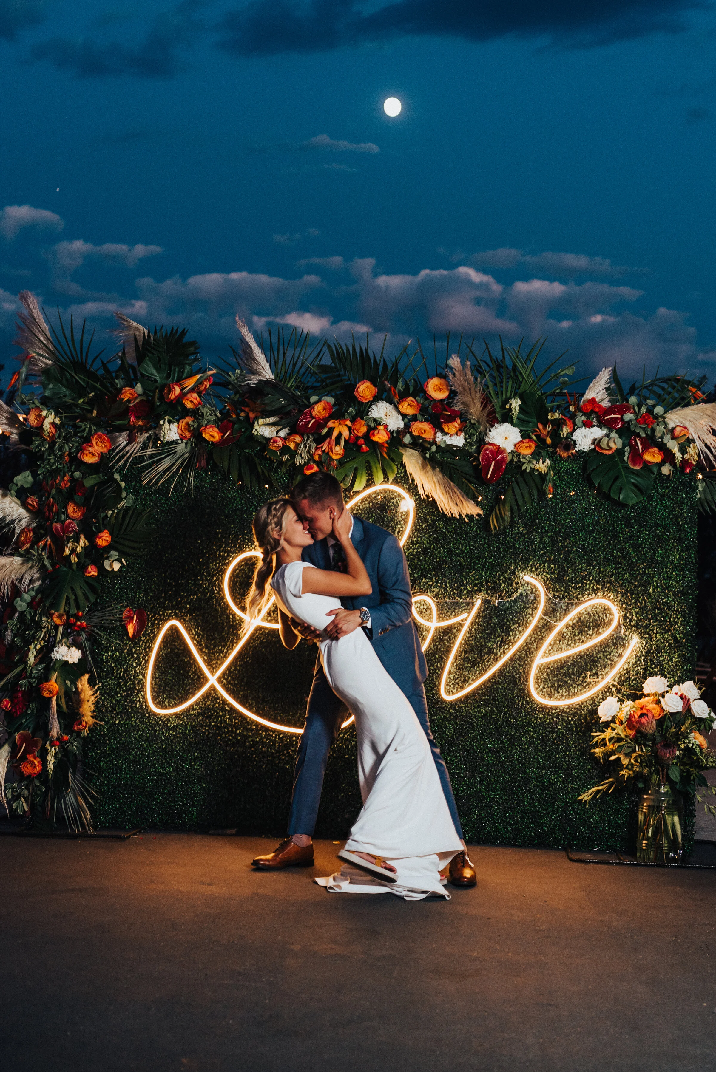  Gorgeous couple in front of neon sign and floral backdrop at this fun-loving backyard wedding reception in Park City. Kristi Alyse Photography Logan Utah photographer Park City wedding photographer Drive-Thru wedding COVID wedding neon signs sociall