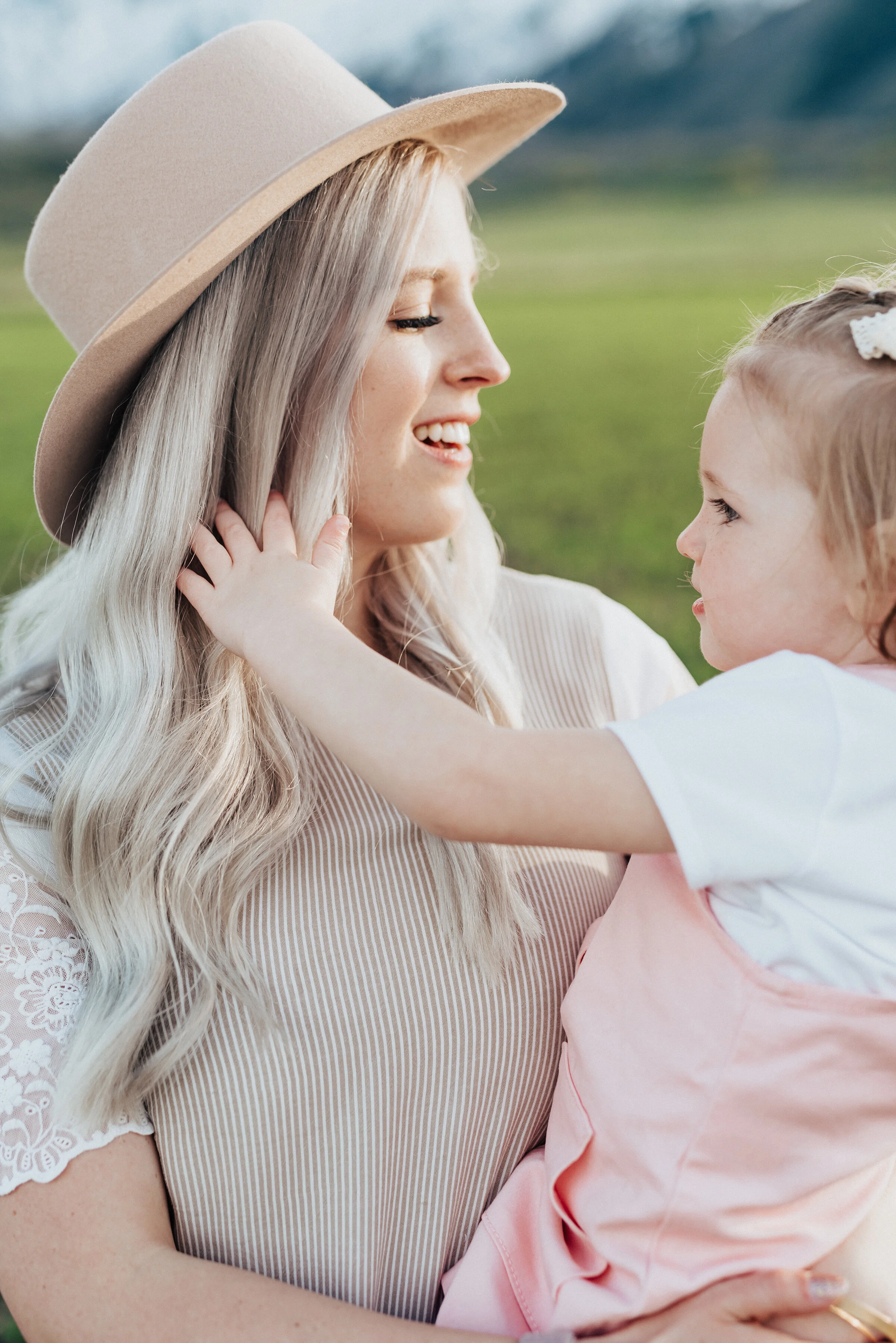  Woman in wide-brimmed hat looks down at precious daughter dressed in pink overalls during a family photography session in logan utah. Mommy and me photography pink outfits for little girls family photography session logan utah photography hipster mo