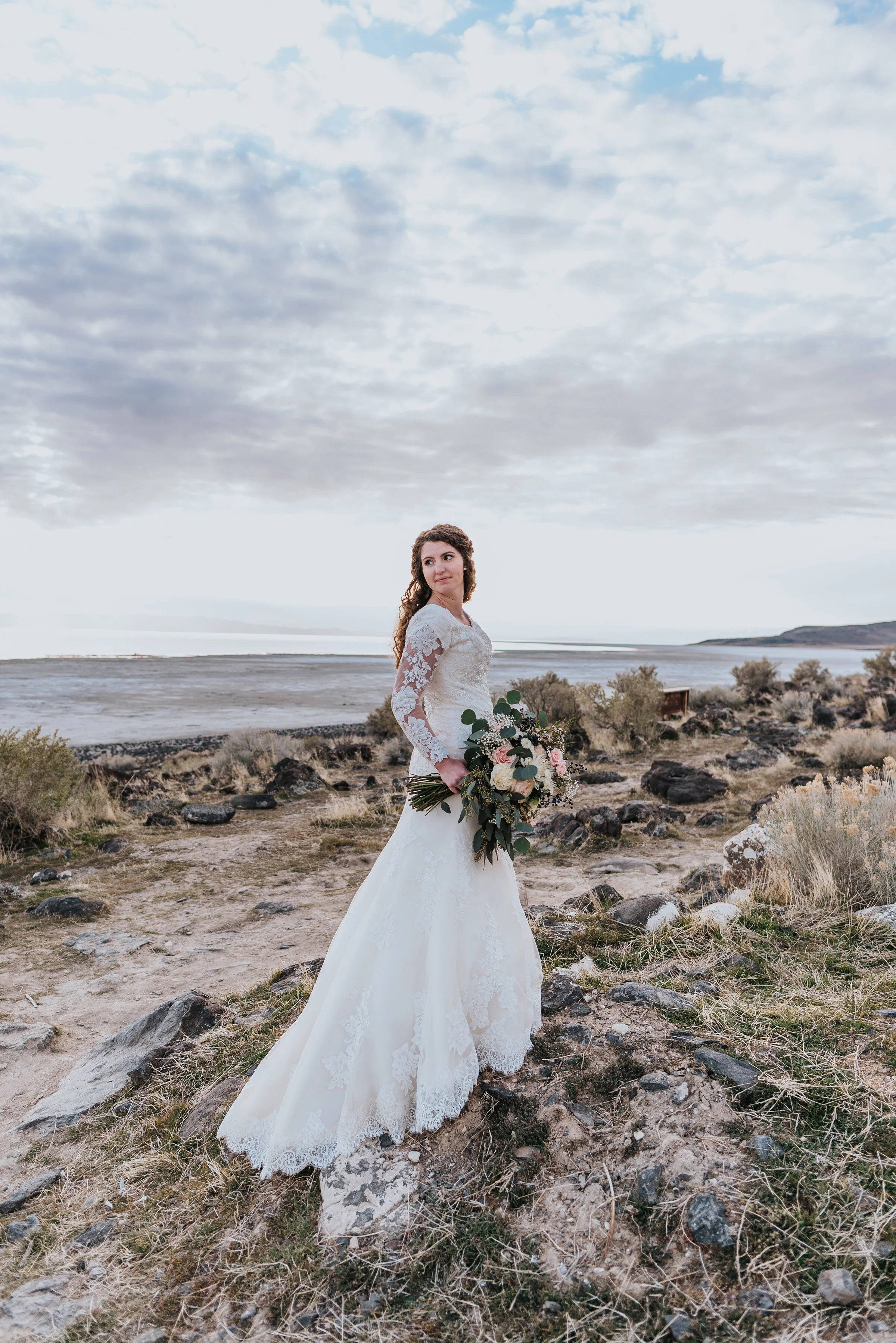  Absolutely stunning bride at the Spiral Jetty in a lace long sleeve wedding gown with the sand and water in the background and the dimension of the rocks at her feet. Wedding formal photoshoot in Spiral Jetty Northern Utah Great Salt Lake photograph