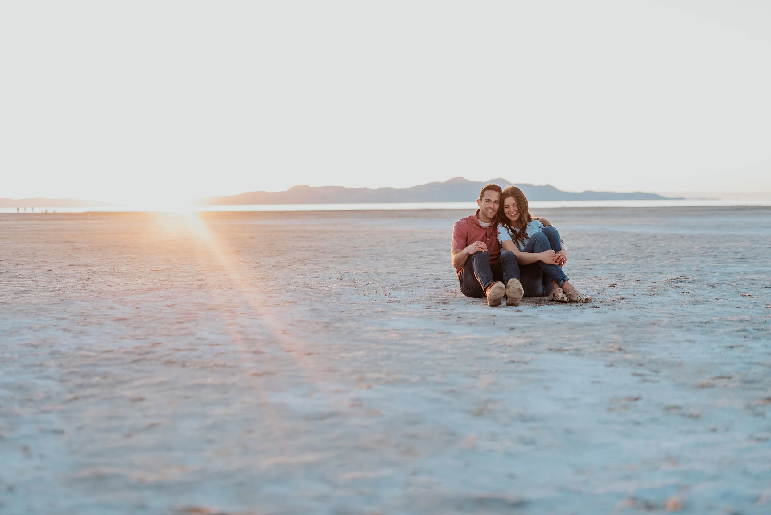 Couple sit together and laugh in saltair and on the salt flats in magna utah during their engagement photoshoot. Outdoor engagement photo ideas using the salt flats for background in photos neutral background photos salt lake city utah engagement ph…