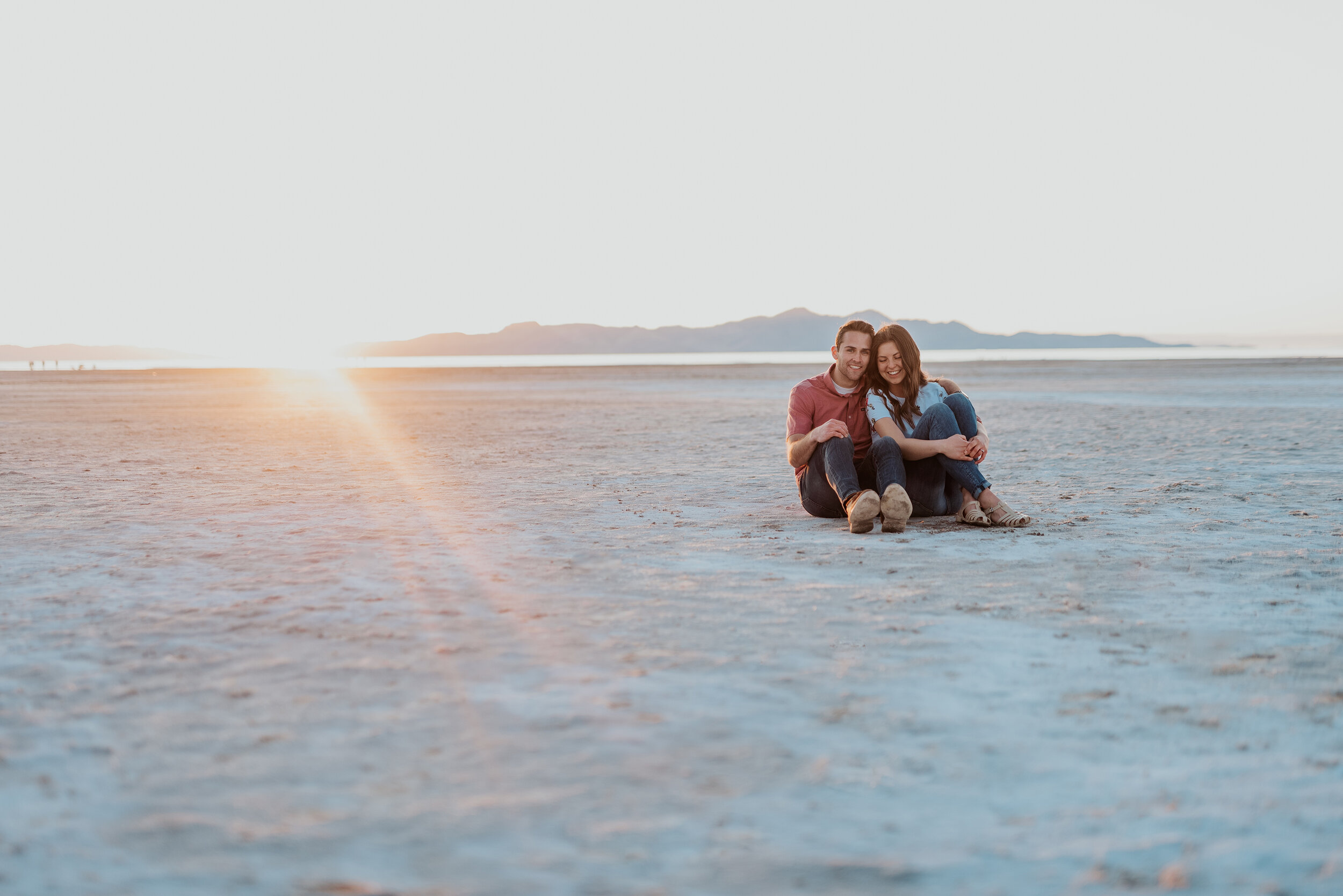 Couple sit together and laugh in saltair and on the salt flats in magna utah during their engagement photoshoot. Outdoor engagement photo ideas using the salt flats for background in photos neutral background photos salt lake city utah engagement photos couple pose by sitting on a dry lake bed #kristialysephotography #kristialyse #utahengagements #engaged #neutralbackground #engagementphotos #coupleposes #magnautah