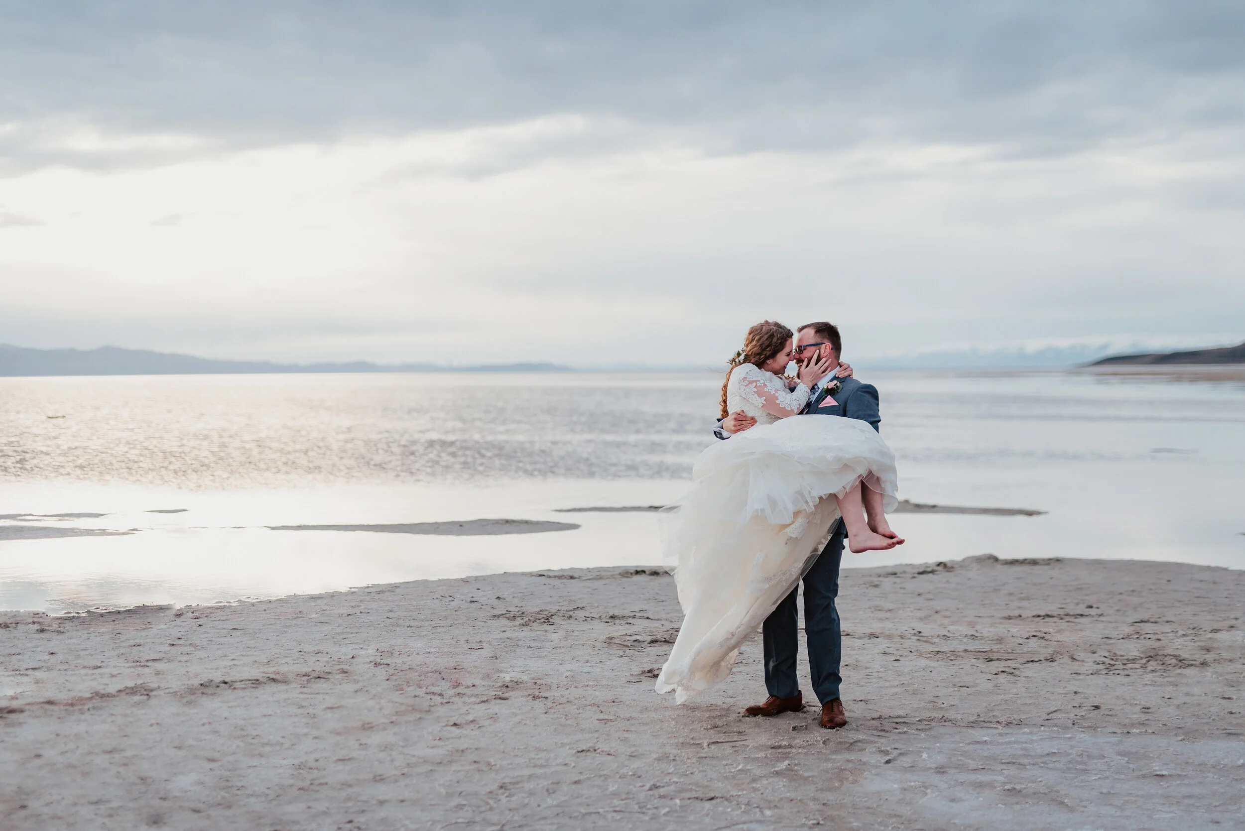 Bride being carried across the sand as her wedding gown blew in the wind at the Spiral Jetty in northern Utah. wedding formal photoshoot in Spiral Jetty Northern Utah Great Salt Lake photography wedding formals natural photo aesthetic bride and groo…