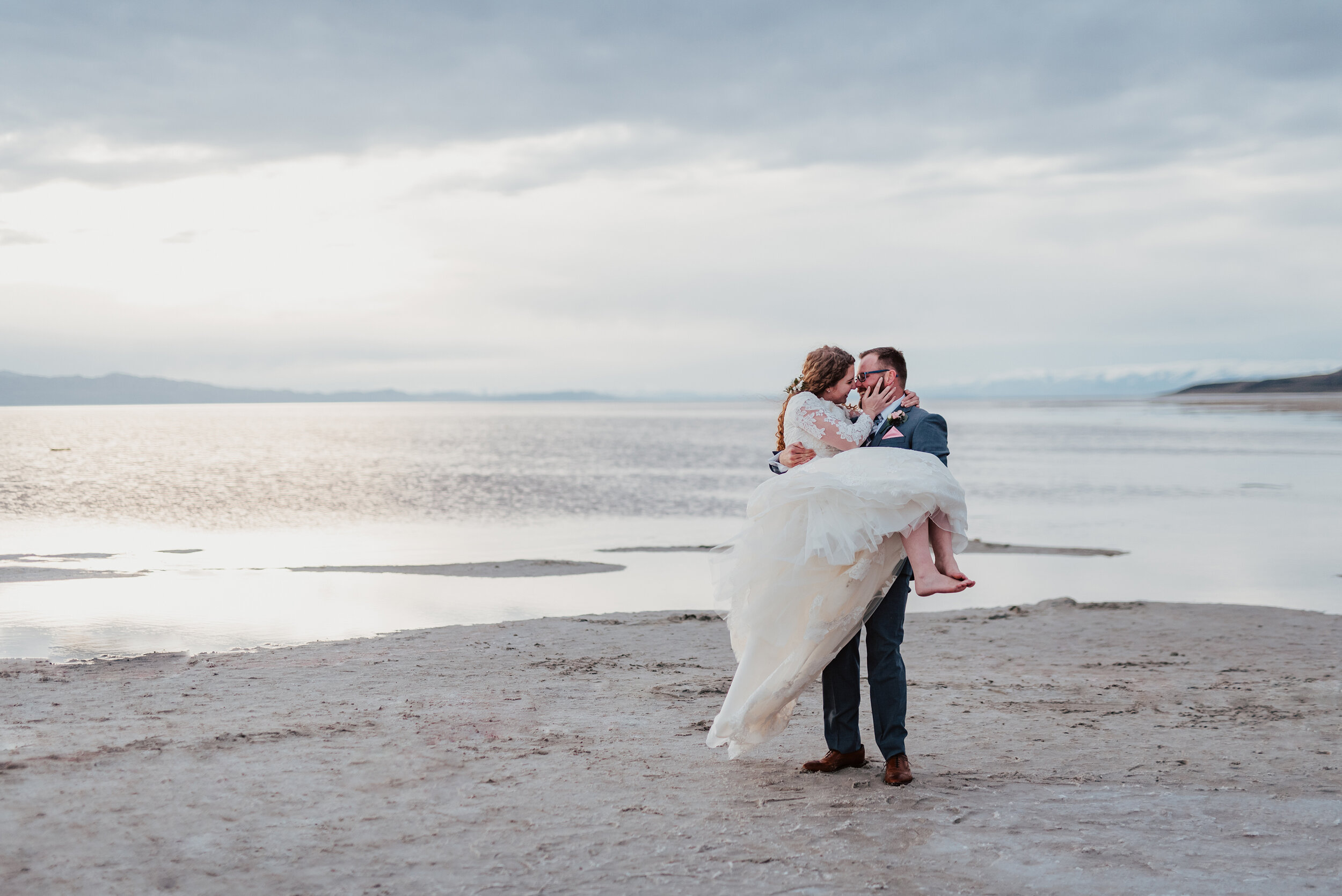 Bride being carried across the sand as her wedding gown blew in the wind at the Spiral Jetty in northern Utah. wedding formal photoshoot in Spiral Jetty Northern Utah Great Salt Lake photography wedding formals natural photo aesthetic bride and groom #spiraljetty #utahphotography #weddingformals #gettingmarried #weddingattire #utahwedding #greatoutdoorswedding #weddingformalsphotoshoot #naturalbeauty