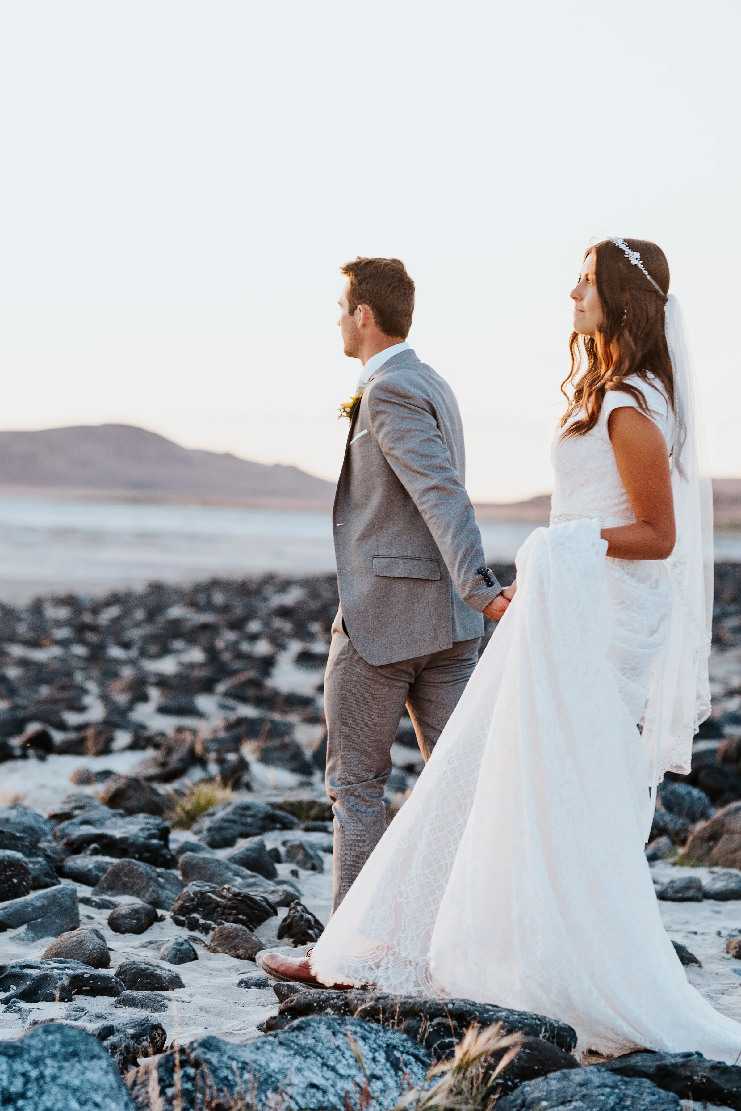  The bride and groom holding hands walking along the rocky shore of the Spiral Jetty in Northern Utah for their wedding formal session. #spiraljetty #GreatSaltLake #formalsession #SaltFlats #sunsetphotosession #NorthernUtah #weddingphotographer #form