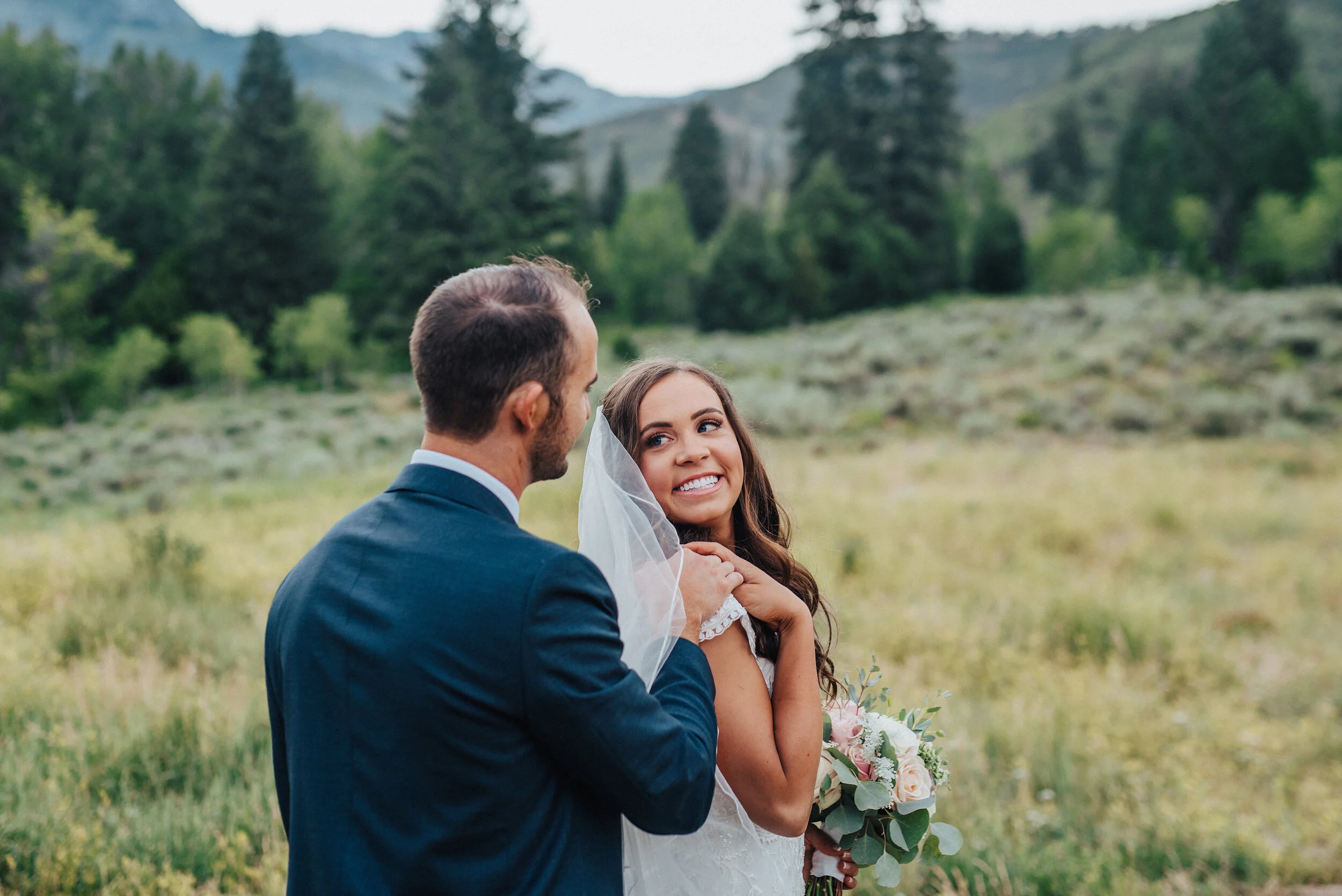 A young bride glances over her shoulder to smile at her new husband while softly touching his hand at Tibble Fork Reservoir. American fork canyon bridal shoot husband and wife navy suit outdoor photoshoot rolling hills woodsy theme evergreen trees a…