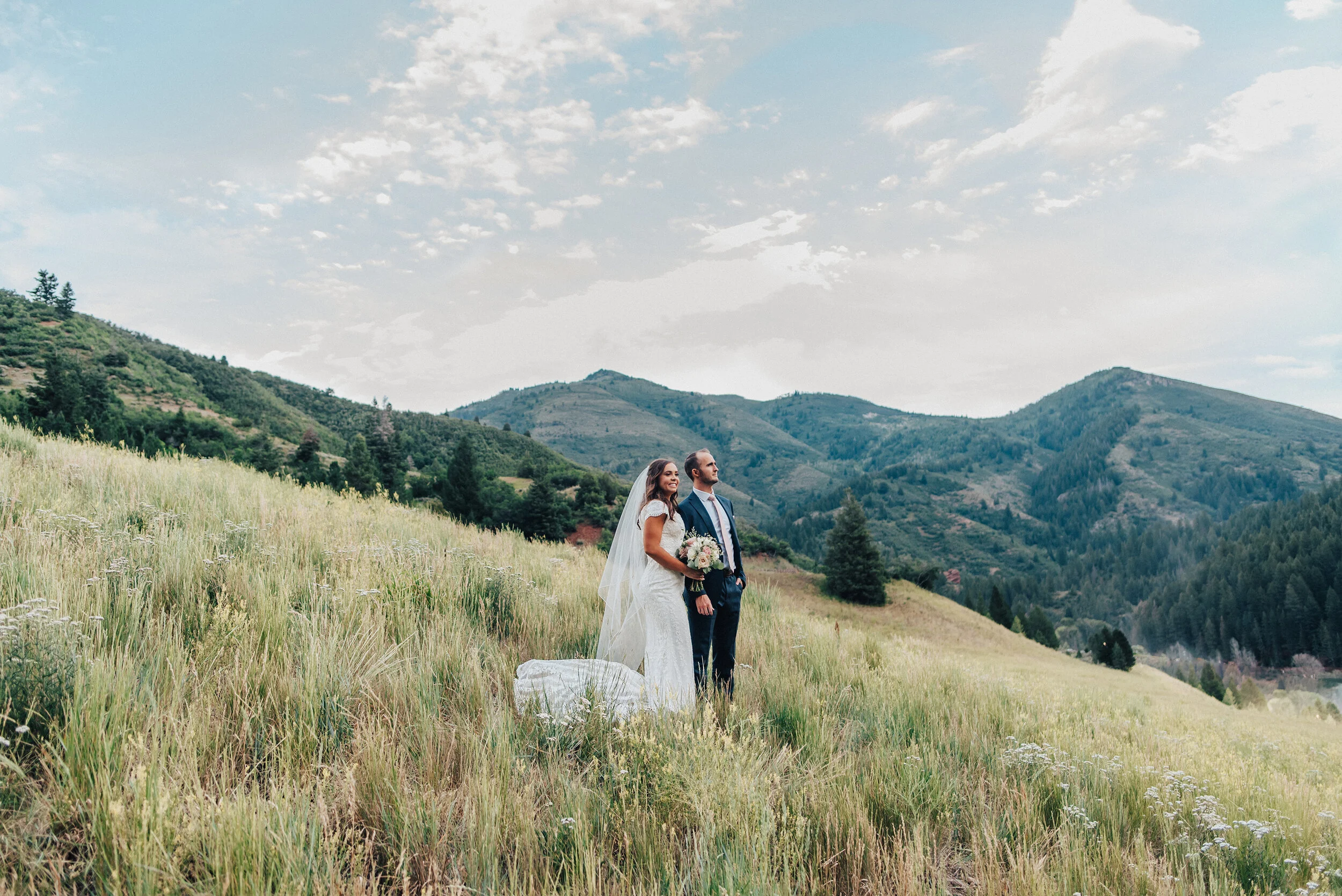 A beautiful couple overlook the Tibble Fork Reservoir while standing on a gorgeous grassy hillside in American Fork Canyon. Mountains in the background evergreen trees couple posed looking forward utah photography wide landscape tall grass navy suit…