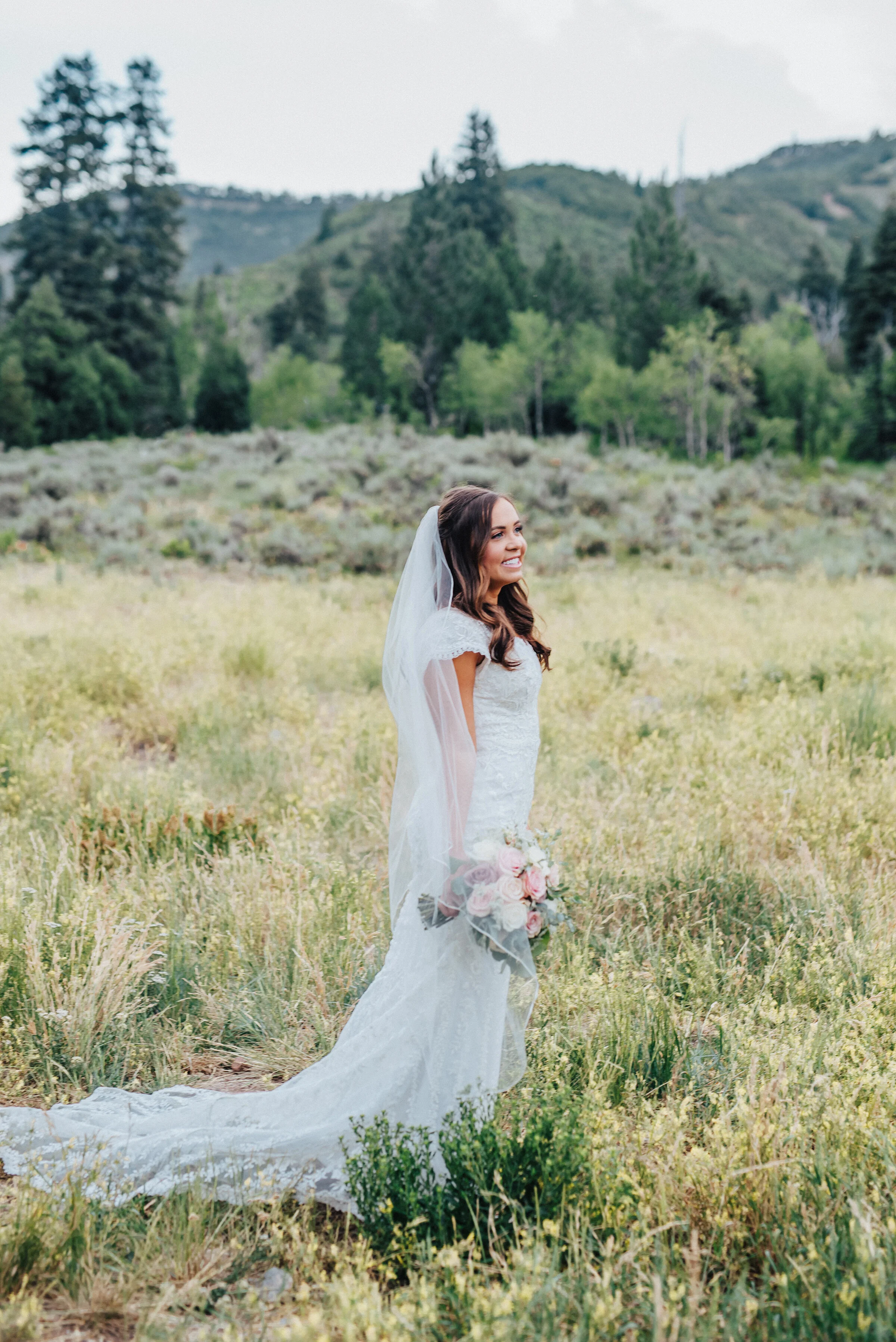  A lovely bride holds her bouquet tightly at her side while smiling and looking across a grassy field in American Fork canyon. Tibble Fork reservoir overlook grassy hillside evergreen trees woodsy scenery bridal shot light pink bouquet mountains in t