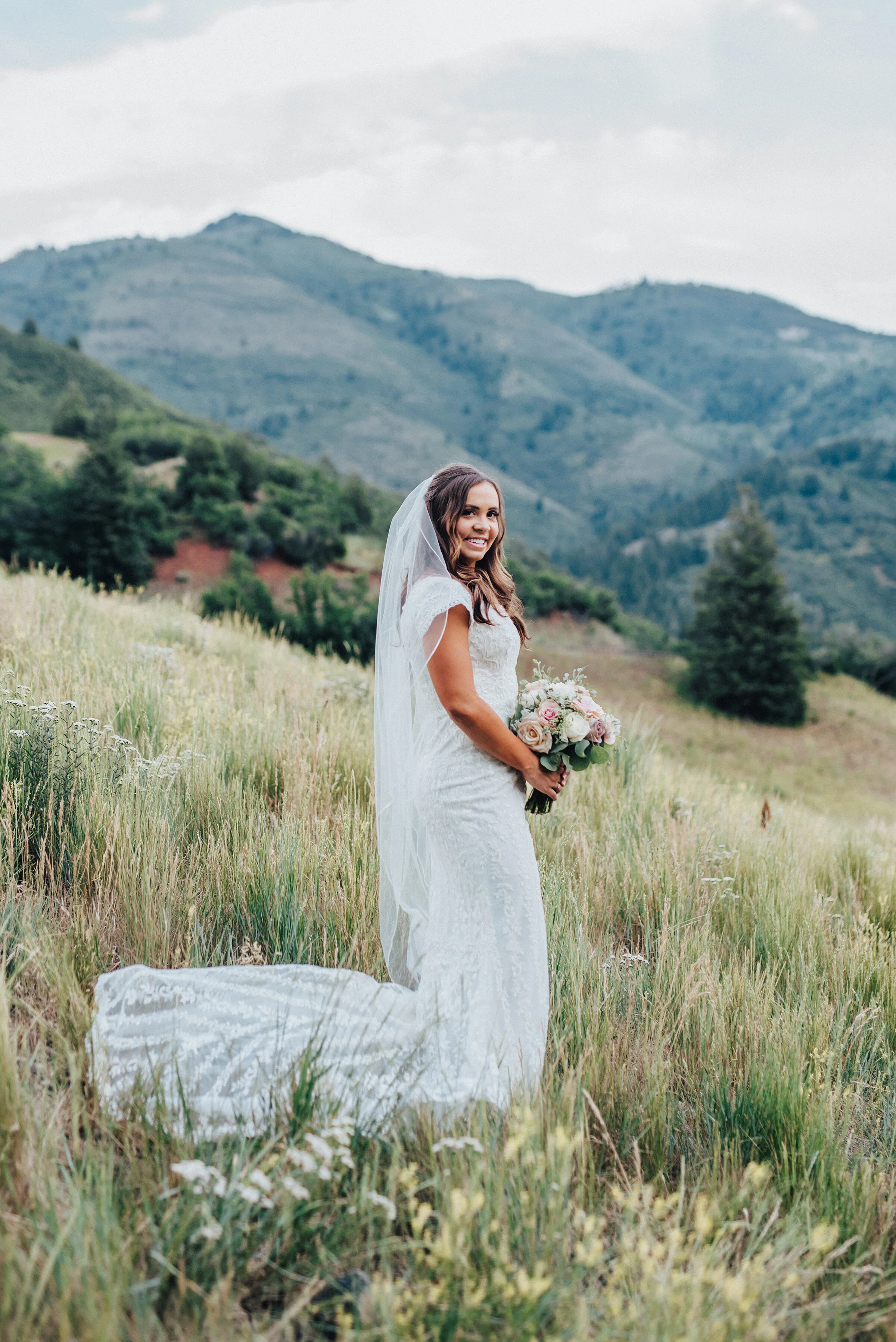  A gorgeous bride holds her bouquet in a traditional position while smiling and looking into the camera on a grassy hillside in American Fork canyon. Tibble Fork reservoir overlook grassy hillside evergreen trees woodsy scenery bridal shot light pink