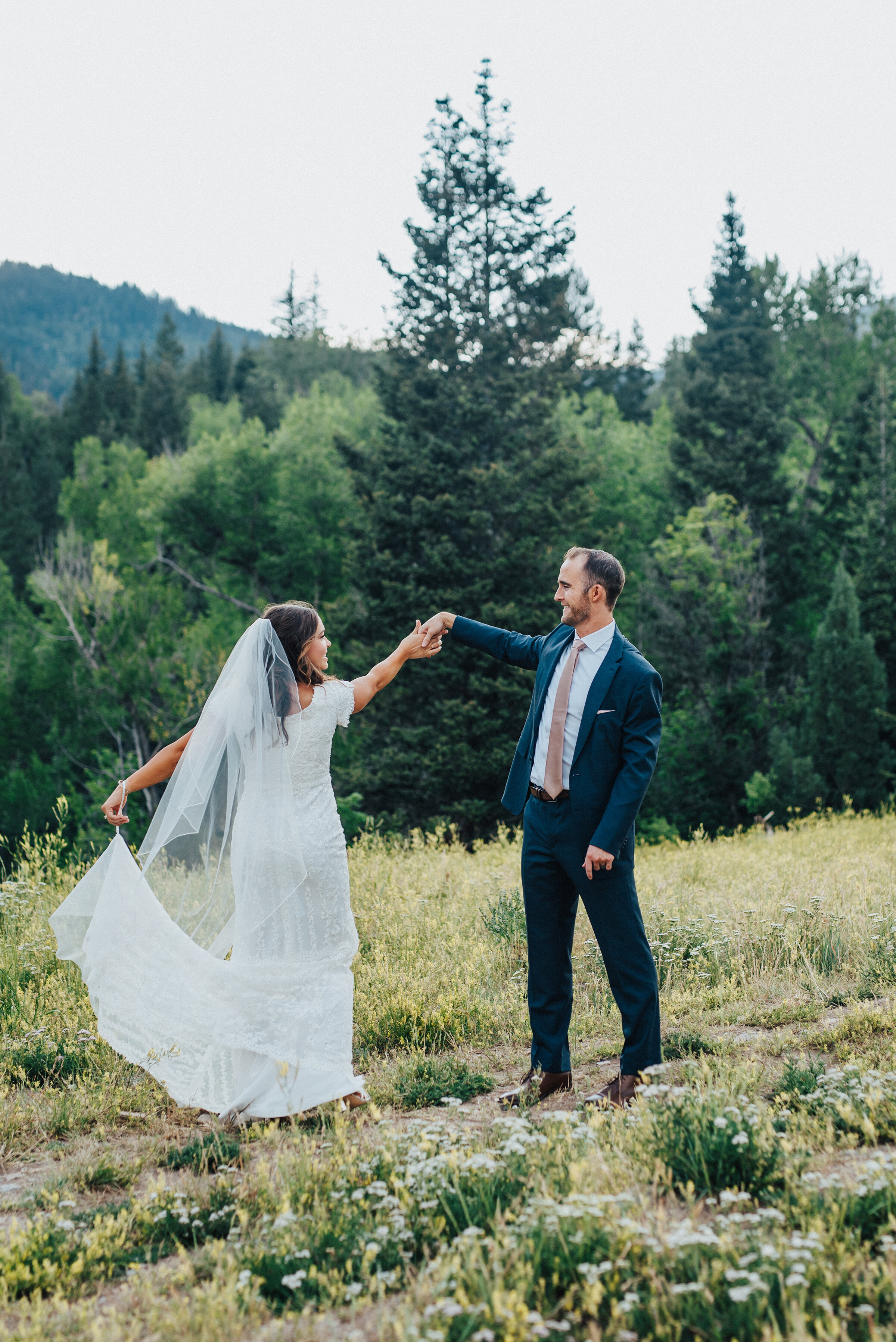  A beautiful smiling couple is captured mid-twirl while the stunning bride lifts her wedding dress just a little bt above the grassy terrain. Mountains in the background evergreen trees couple looking at each other utah photography landscape twirling