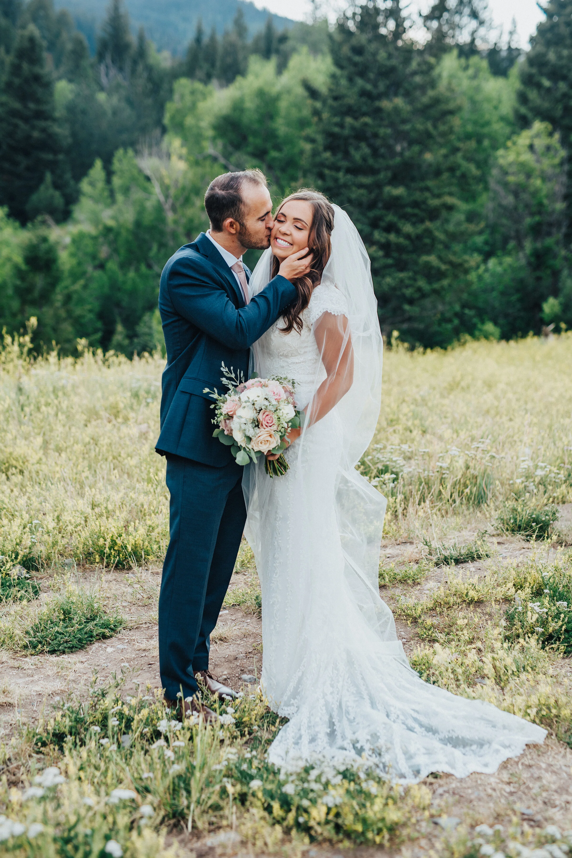  A smitten husband gently caresses his smiling bride’s cheek while giving her a soft kiss.holding bouquet in traditional position evergreen trees eyes closed utah photography landscape navy blue suit light pink tie light pink bouquet #husbandandwife 