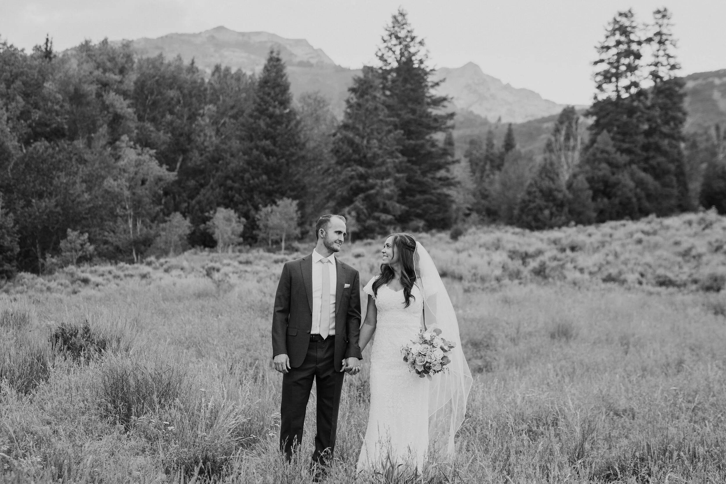 A classic black and white shot of a lovely couple looking into each other’s eyes and smiling while strolling in tall grass. Holding hands walking through the grass evergreen trees black and white photography exchanged looks wide landscape #husbandan…