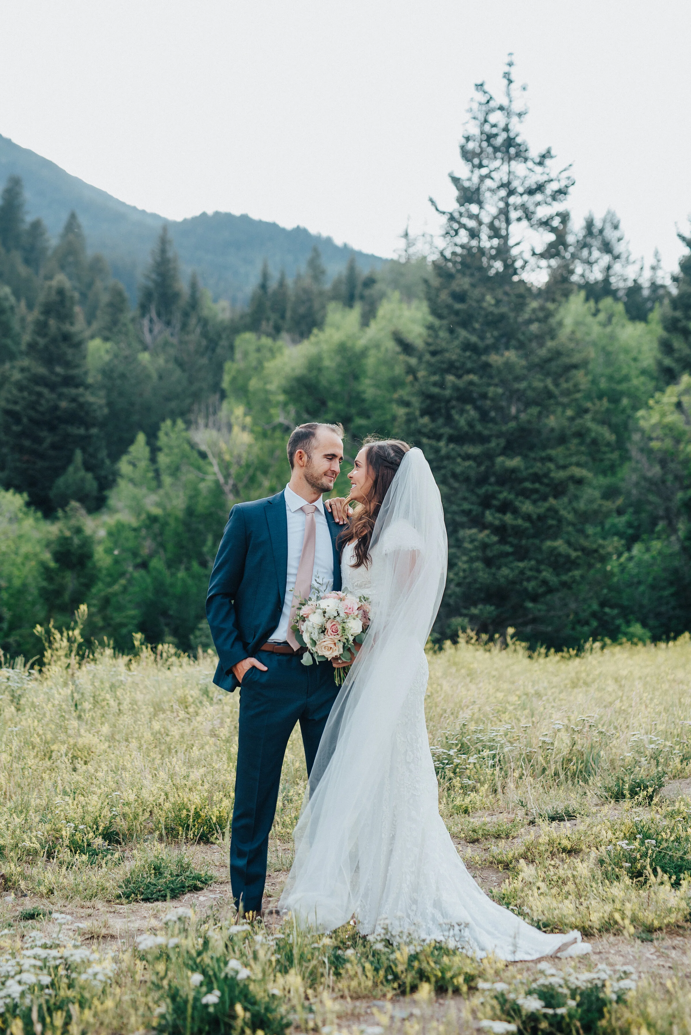  An enamored couple looks at each other while standing in an open grassy area before the woods.Light pink bouquet and tie navy blue suit evergreen trees mountainside background grassy terrain American Fork Canyon Tibble Fork Reservoir full length vei