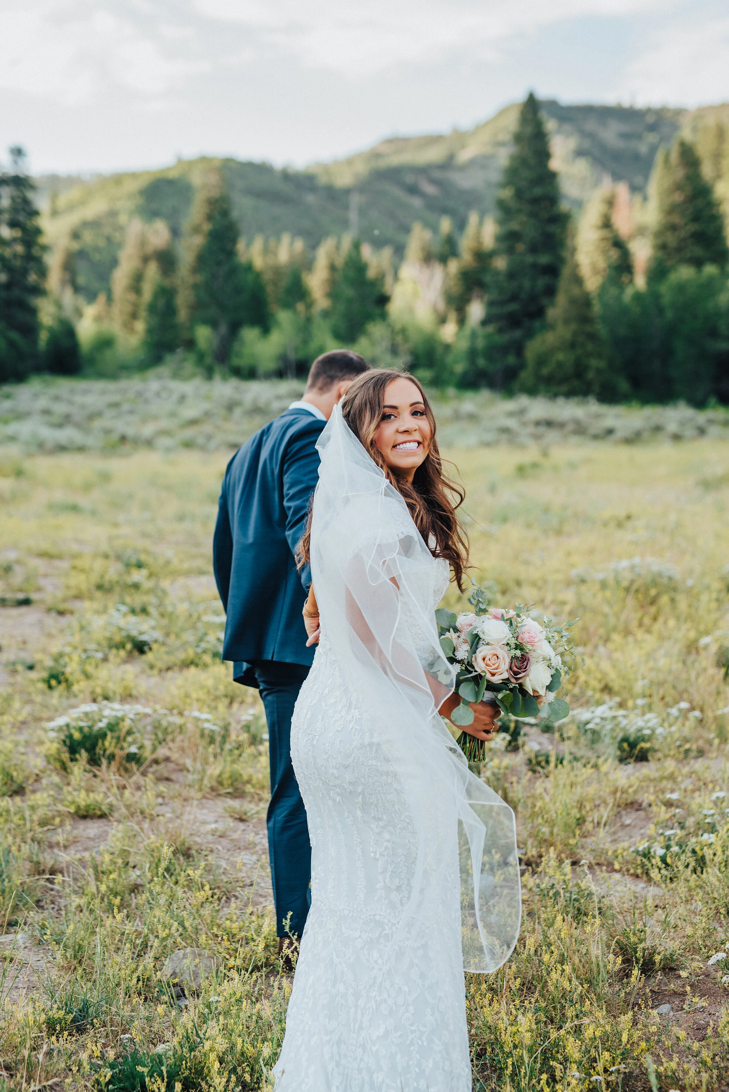  A smiling bride looks behind her as her husband gently tugs her forward towards the beautiful mountainside in American Fork Canyon. Light pink bouquet navy suit strolling in the grass evergreen trees husband and wife holding hands mountains and ever