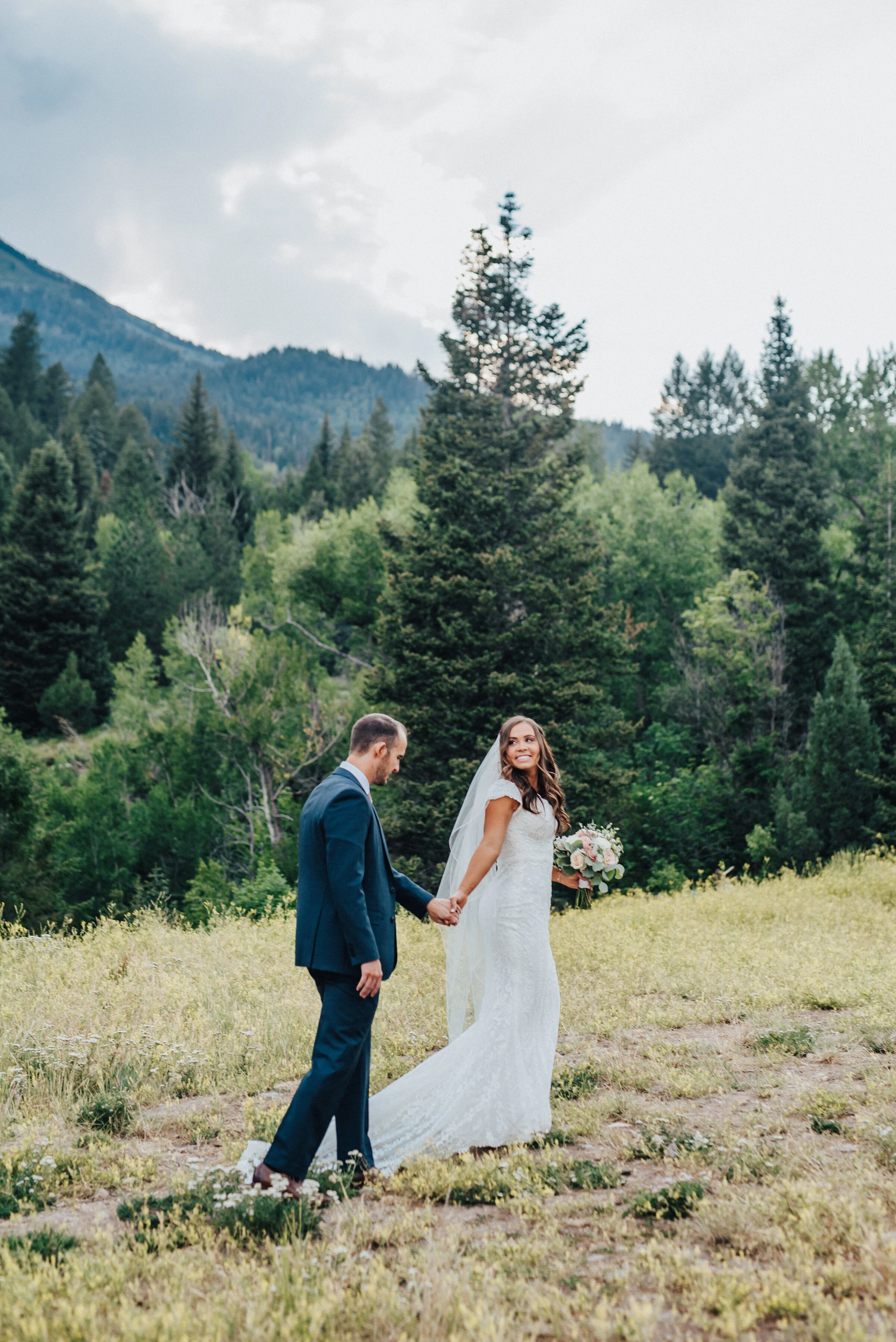 A smiling bride leads her husband by the hand as she strolls across an open grassy area in American Fork Canyon. Light pink bouquet and tie navy suit evergreen trees mountainside background grassy terrain American Fork Canyon Tibble Fork Reservoir h