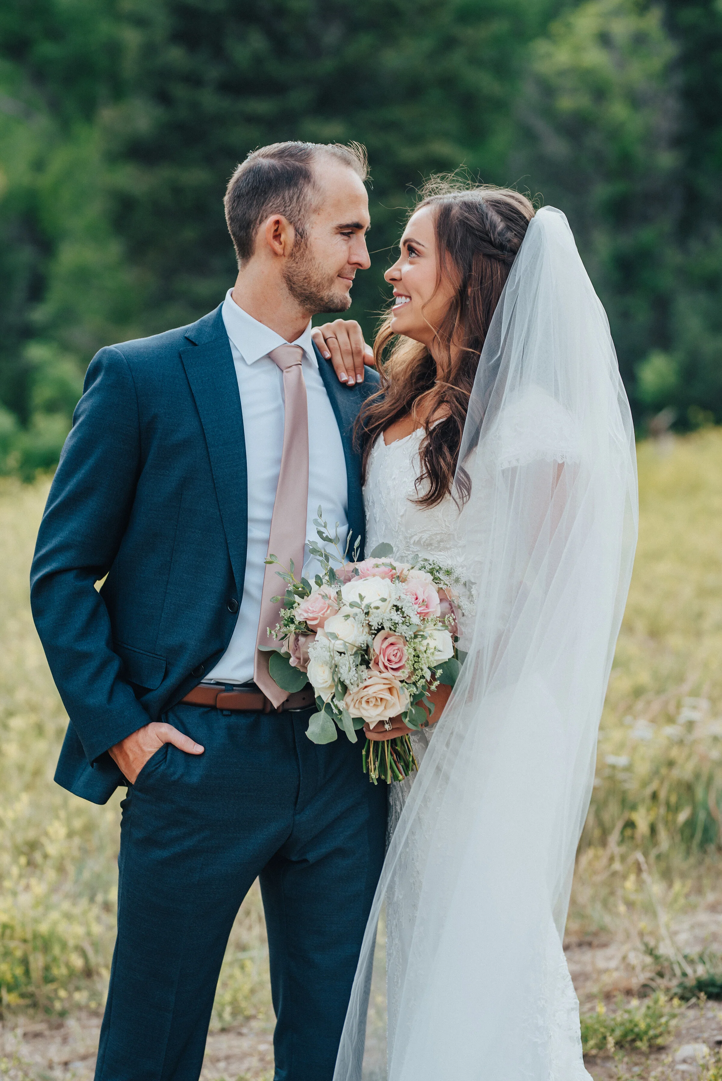 An infatuated husband is looking into his smiling bride’s eyes while she has one hand gently perched on his shoulder and the other is tightly holding her bouquet. Close-up shot full-length veil evergreen trees couple looking at each other utah photo