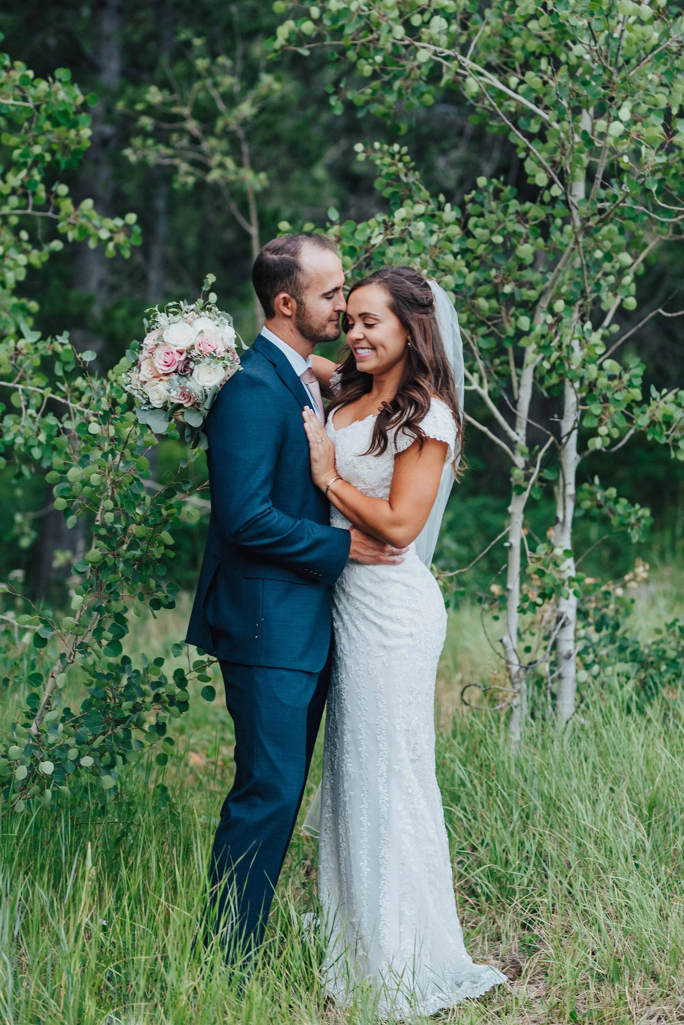  A gorgeous bride smiles as her new husband gently grabs her waist and pulls her towards him while surrounded by beautiful greenery. Close-up shot full-length veil evergreen trees couple looking at each other utah photography landscape navy suit ligh